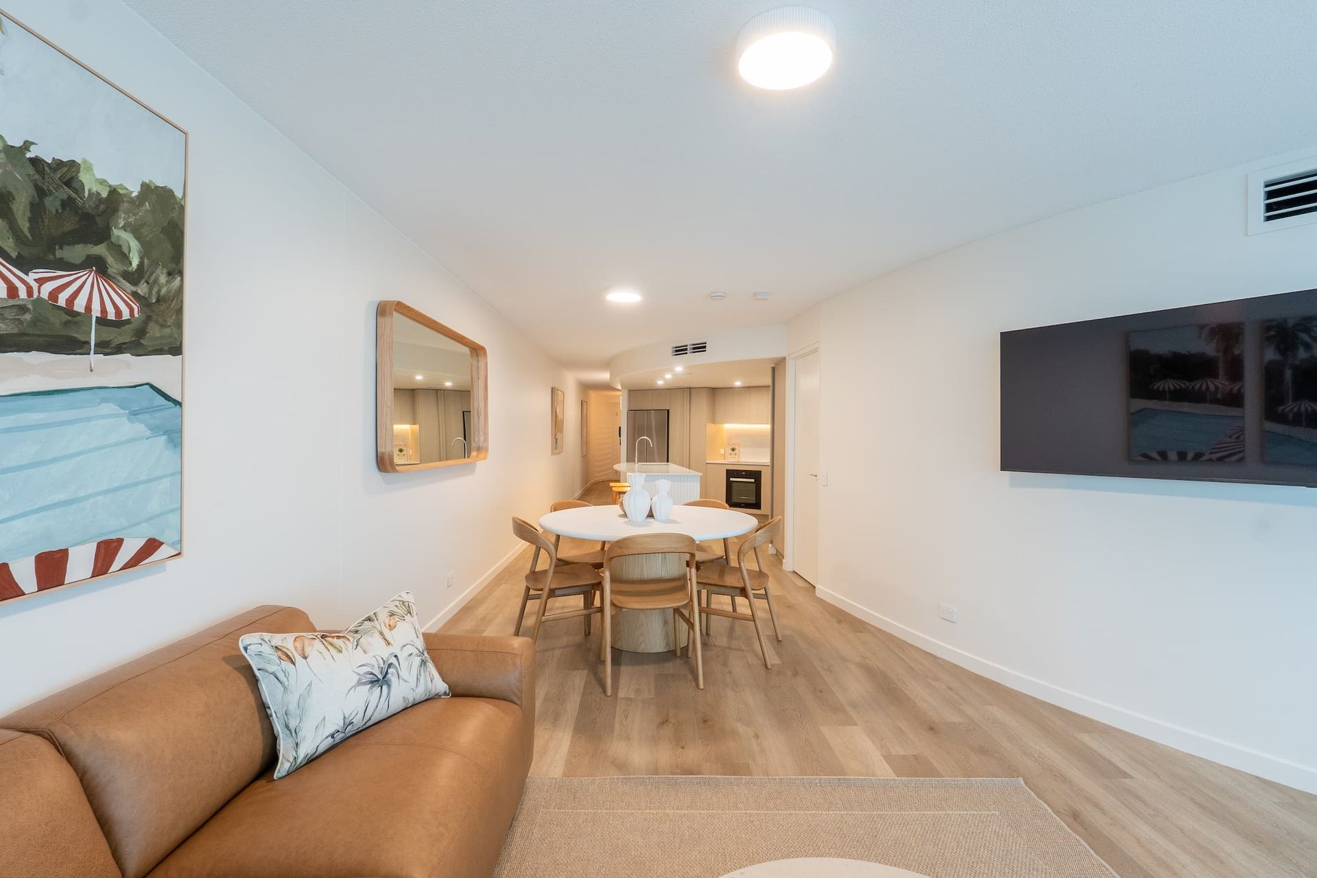 Bright modern living room with tan sofa, wall-mounted TV, and dining table leading to kitchen