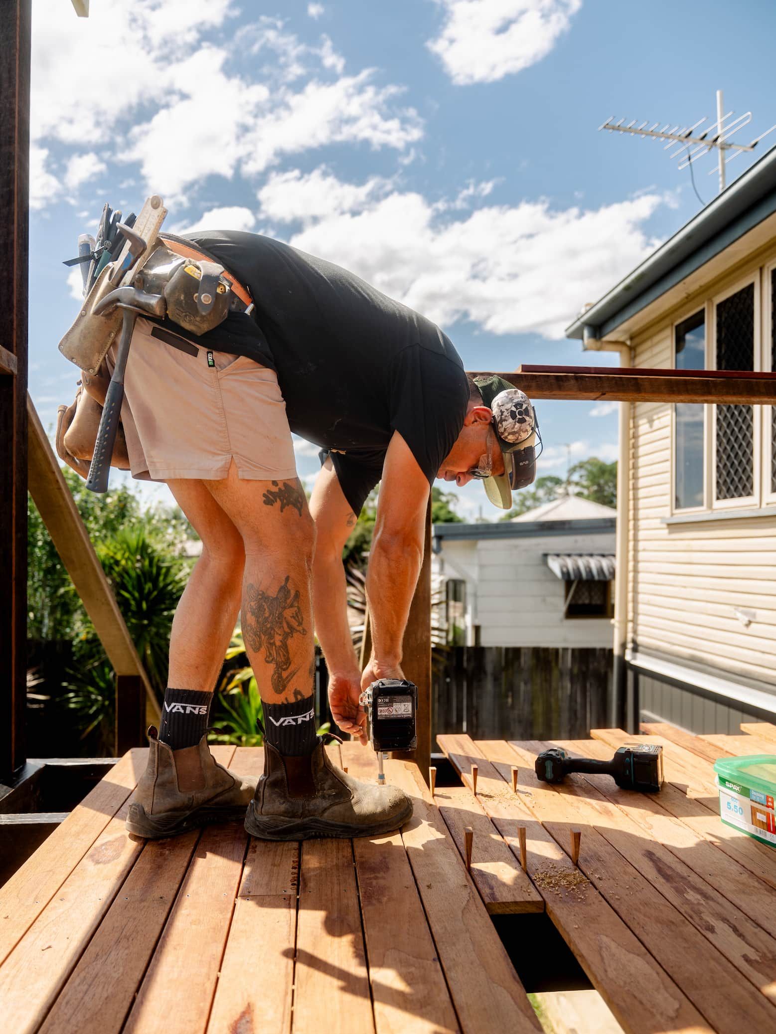 Person bending over on a wooden deck, working with tools beside a house under a bright blue sky.