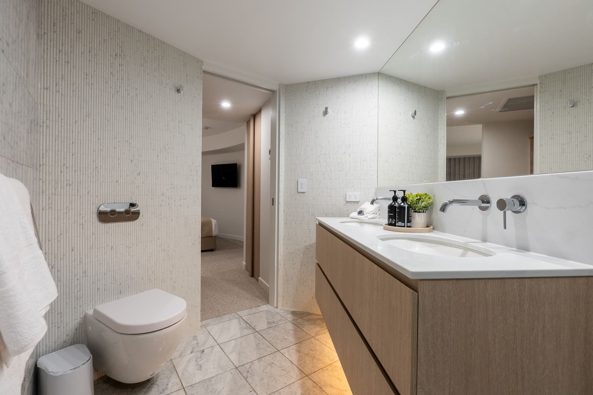 Modern bathroom with double vanity, round mirror, toilet, and beige tile walls and floor.