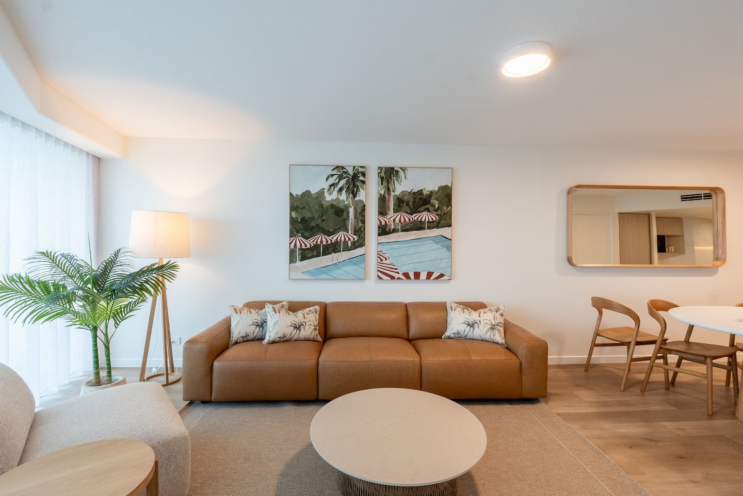 Bright living room with tan sofa, round coffee table, floor lamp, wall art, and dining area by large windows