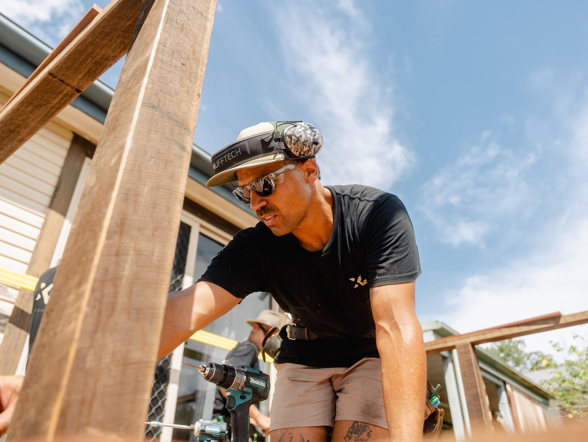 Worker in a cap and sunglasses using a drill on a wooden structure under a blue sky