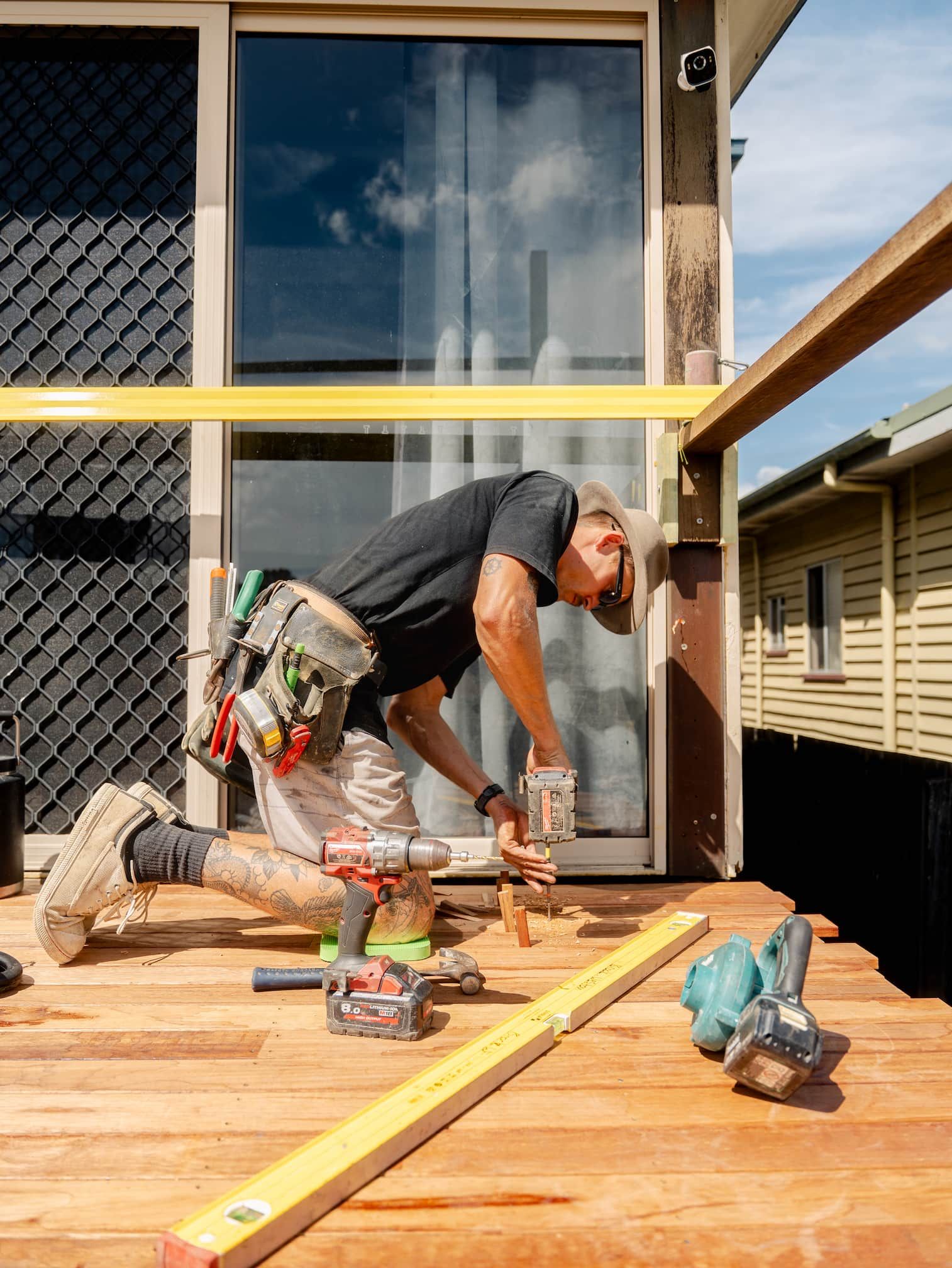 Worker kneeling on a wooden deck, using tools to install a floor near a sliding glass door.