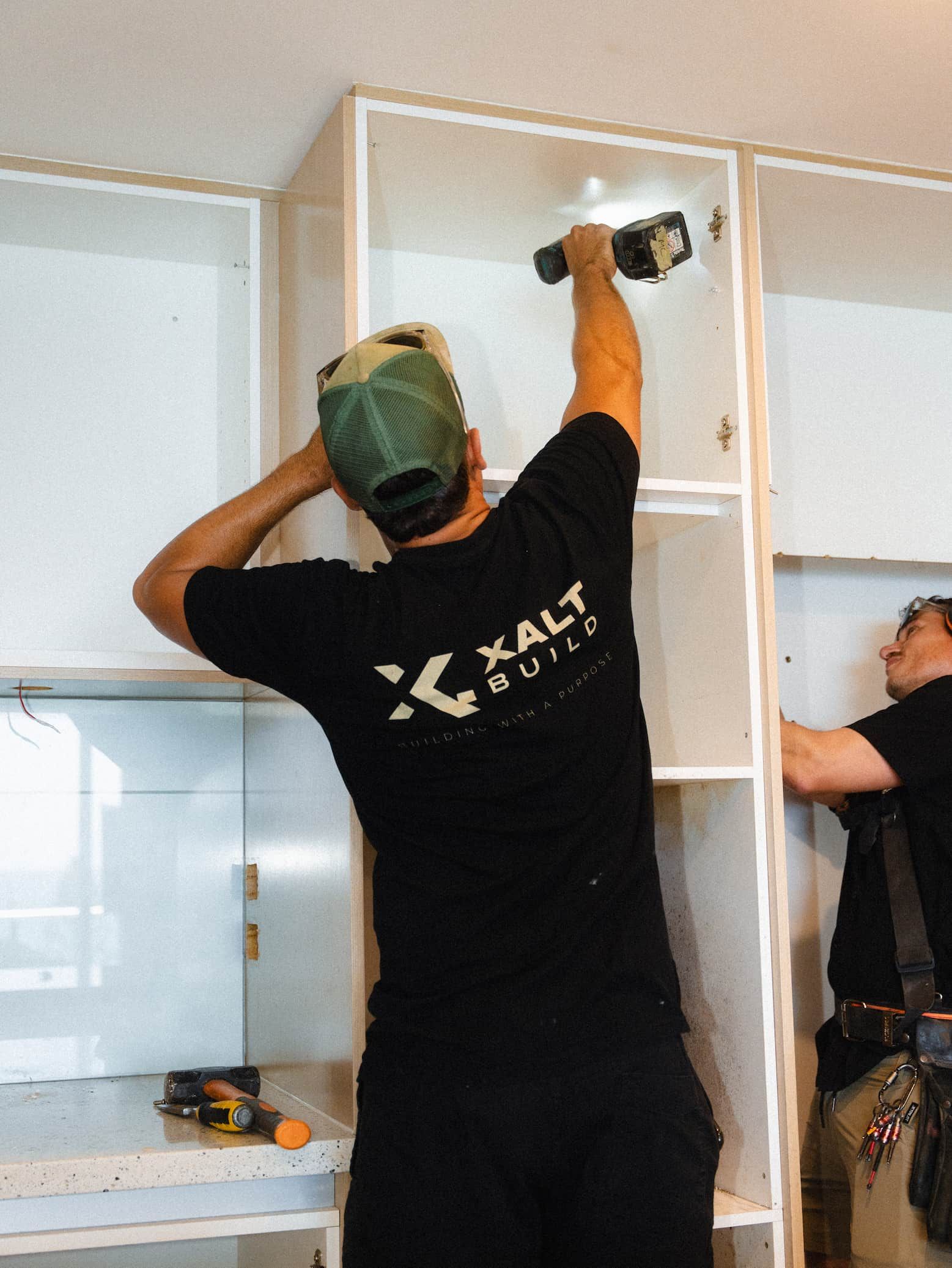 Two workers installing white cabinets in a kitchen, one using a power drill.