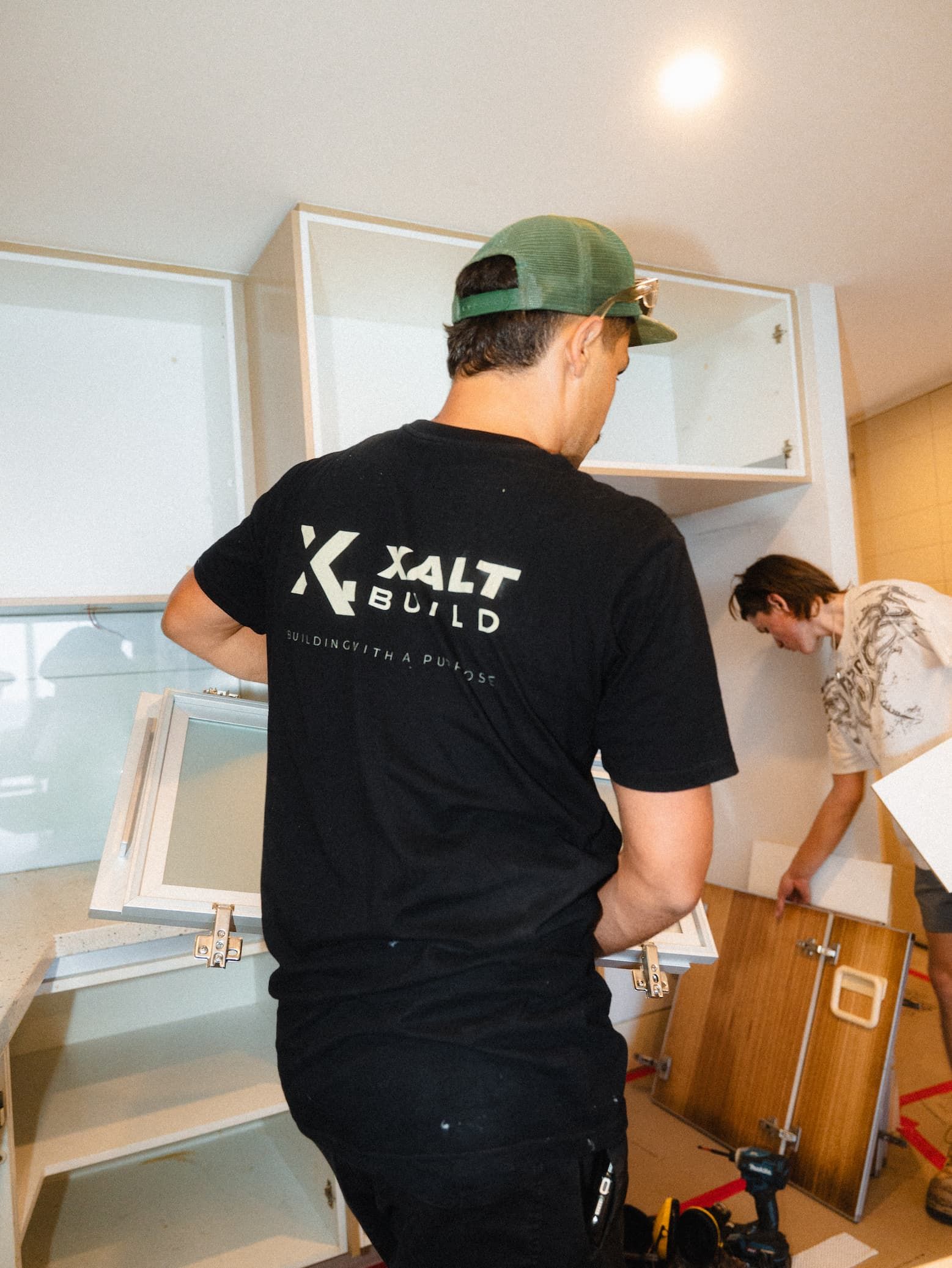 Two people unpacking cardboard boxes in a bright room; one wears a black shirt and green cap.