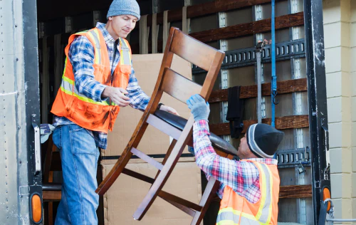 Two movers in safety vests load a wooden chair into a truck.