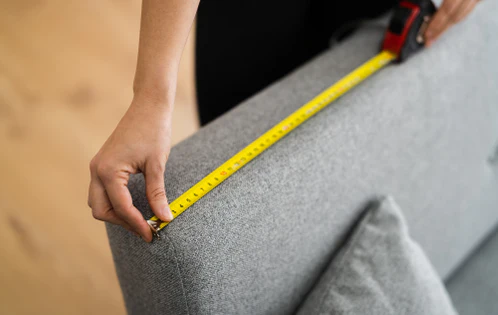 Person measuring grey sofa with yellow tape measure.