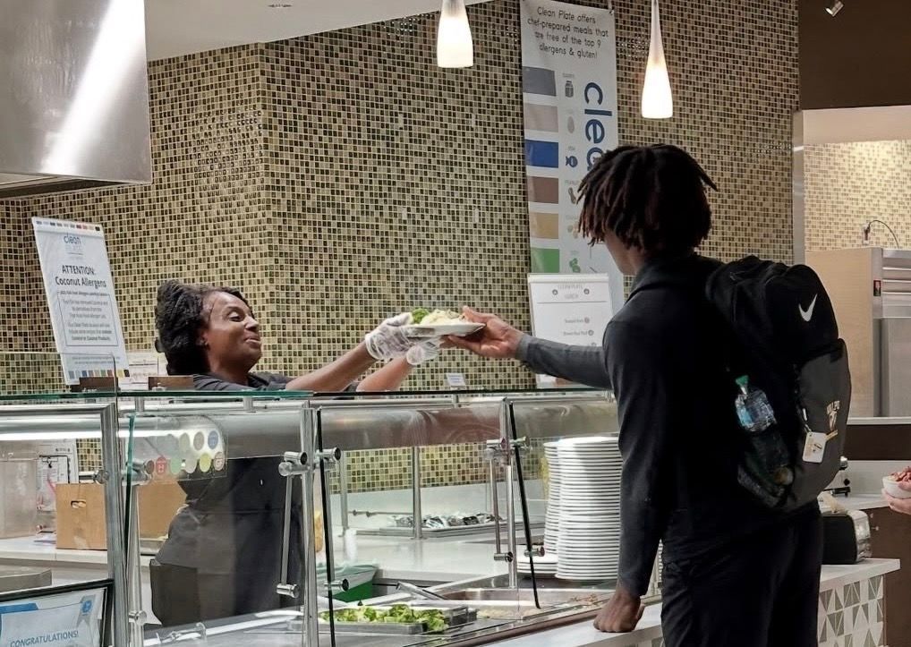 Three people behind a food counter: one with glasses, one in a chef's hat, and one smiling.