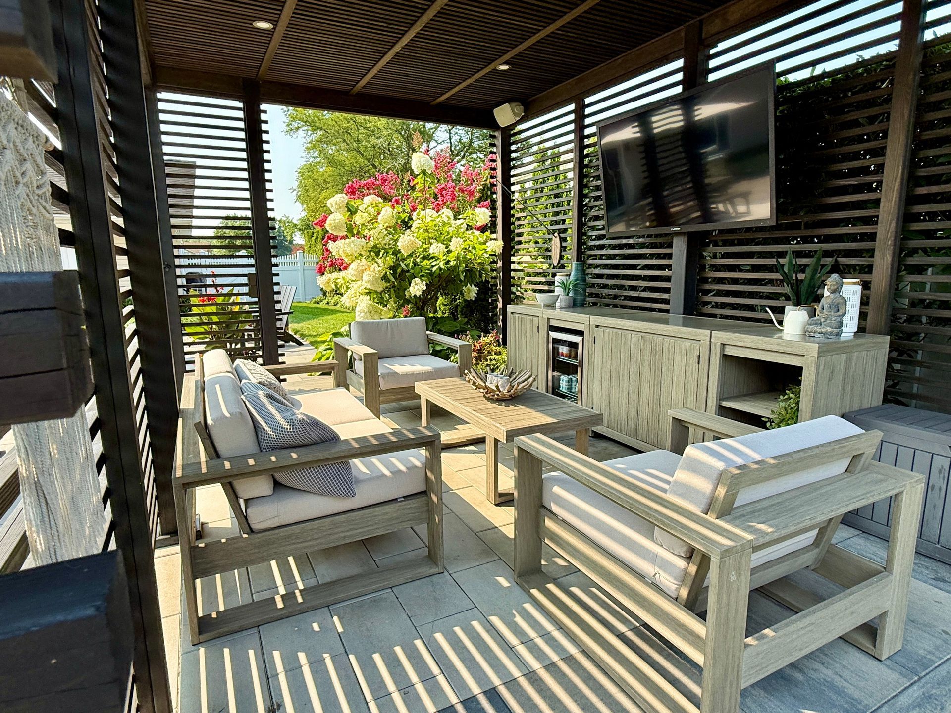 Outdoor lounge area with wooden furniture under a pergola, including a TV and beverage cooler.