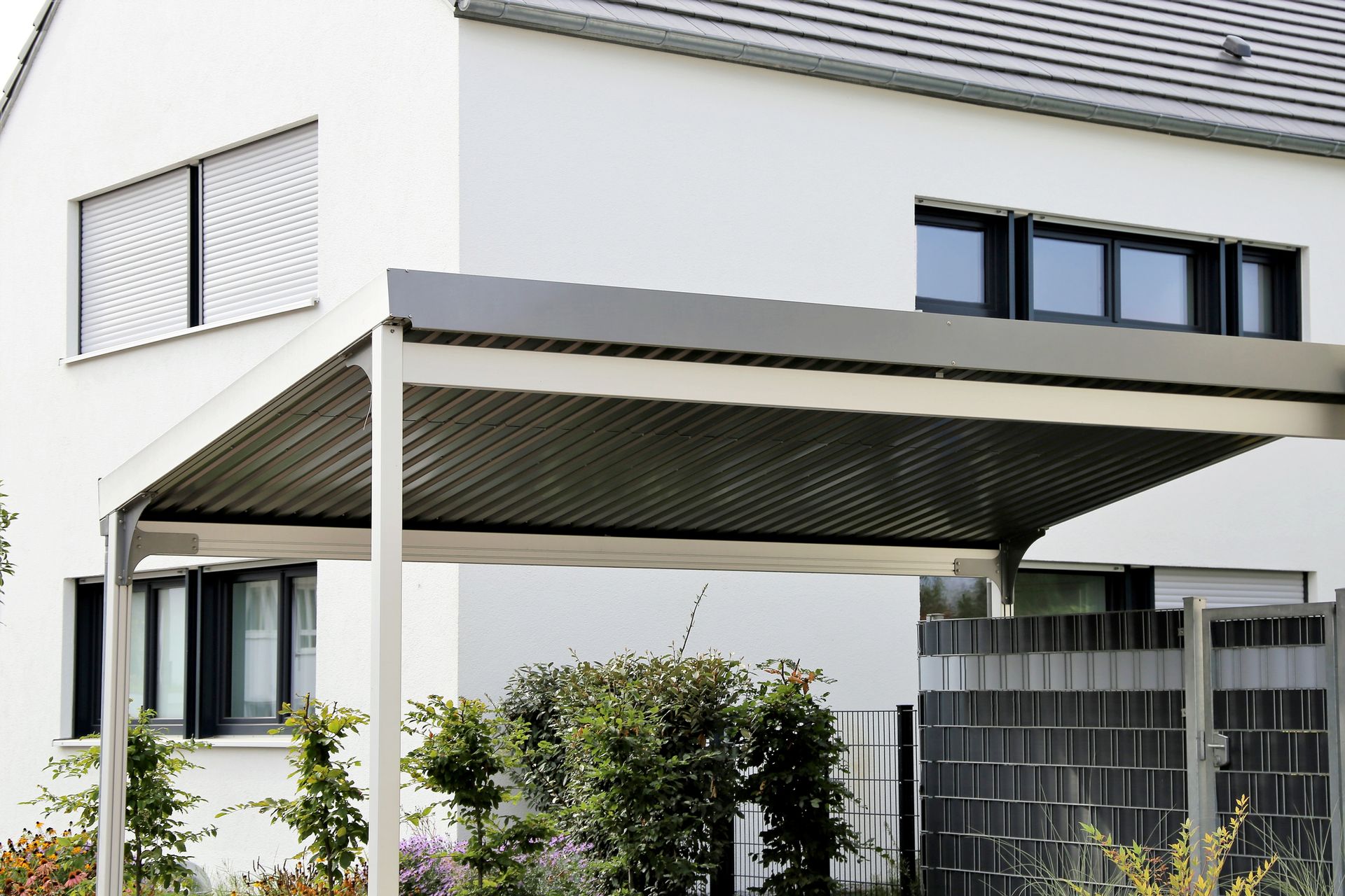 Carport attached to a white house; gray roof, support beams, and window frames. Carport attached to a white house, with a dark roof and steel frame.