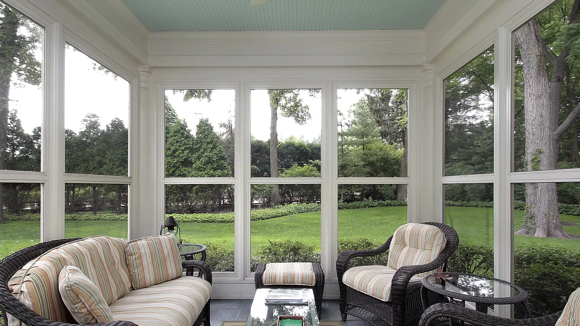 Sunroom with wicker furniture and views of a green yard through large windows.