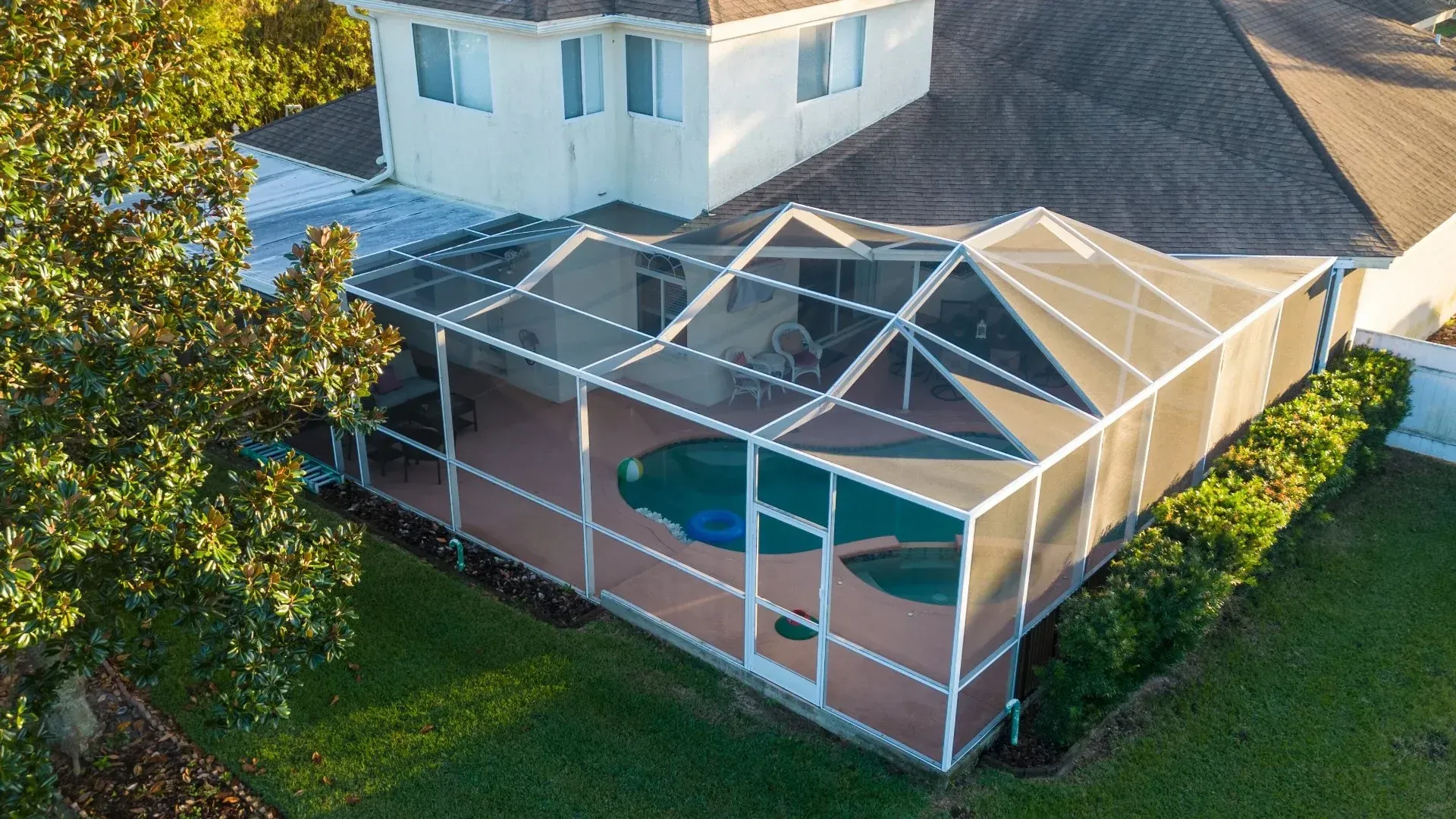 A screened-in pool enclosure attached to a light-colored house with a dark roof and surrounding greenery.