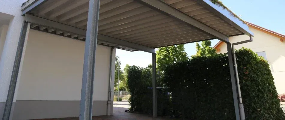 Covered walkway with gray supports and roof, leading to a green hedge and building.