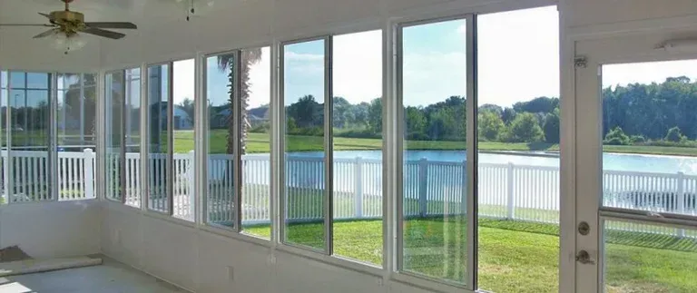 Sunroom with large windows overlooking a lake and white fence. Ceiling fan, green grass, and blue sky.