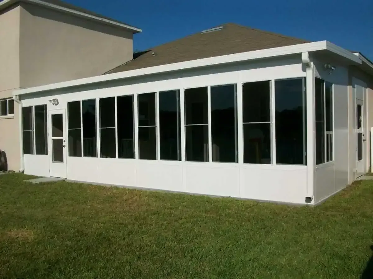 White-framed sunroom with dark windows attached to a beige house, set on green grass under a clear blue sky.