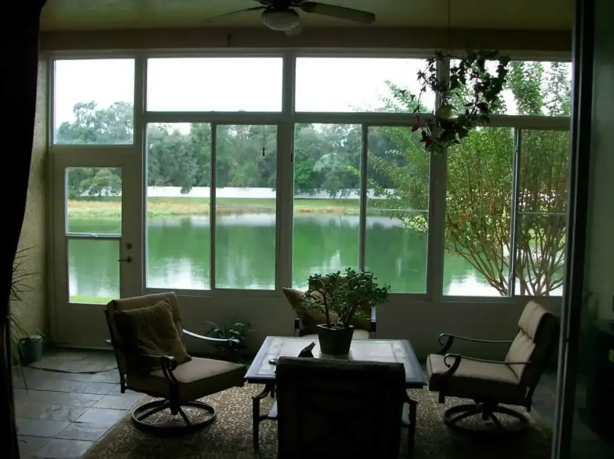 Sunroom with view of a pond, with table and chairs. Light-filled room with large windows, and green landscape.