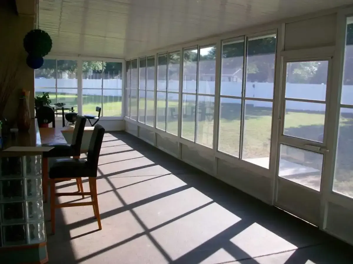 Sunroom interior with a bar and stools, looking out onto a backyard. Sunlight streams through glass windows.