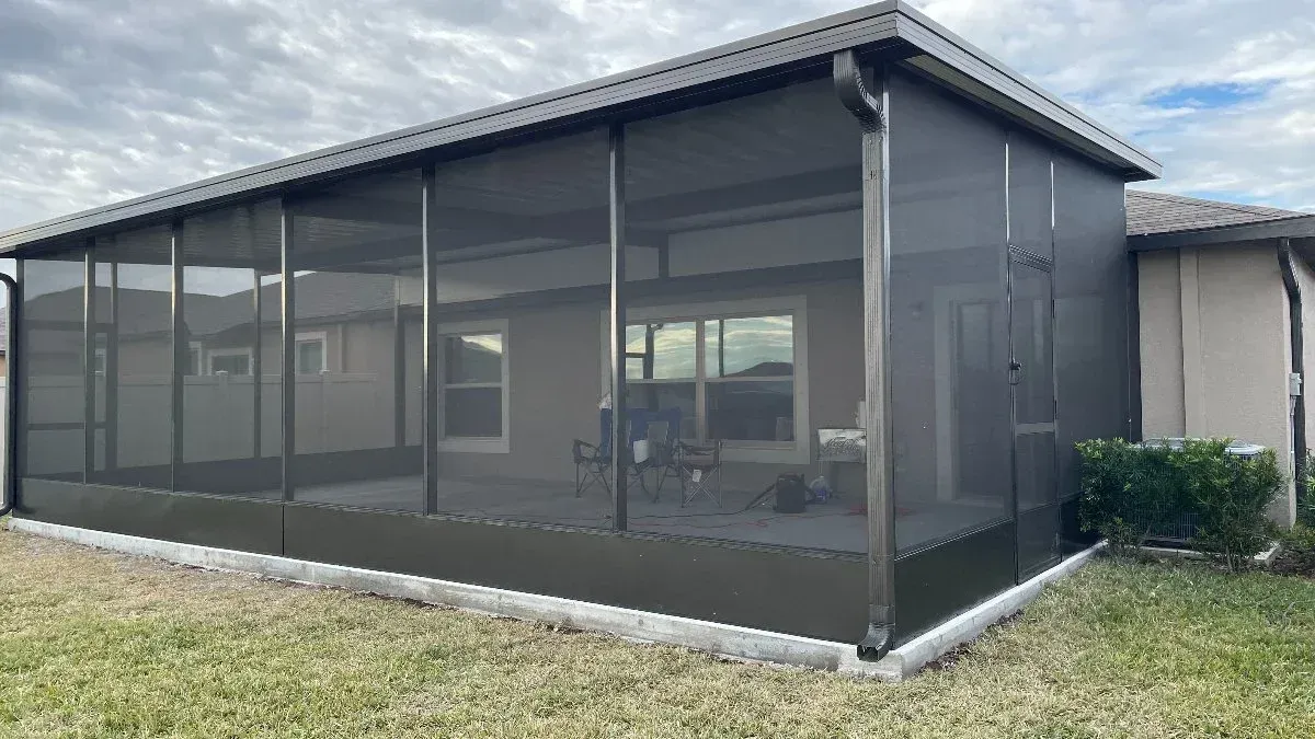 Screened patio addition attached to a light beige house with dark brown trim, gray screens, and a cloudy sky.