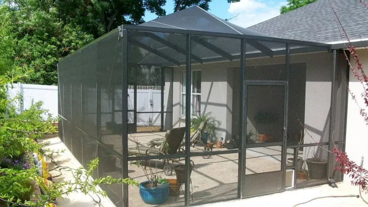 Screened patio enclosure with black frames, filled with plants and patio furniture, attached to a house with a gray roof.