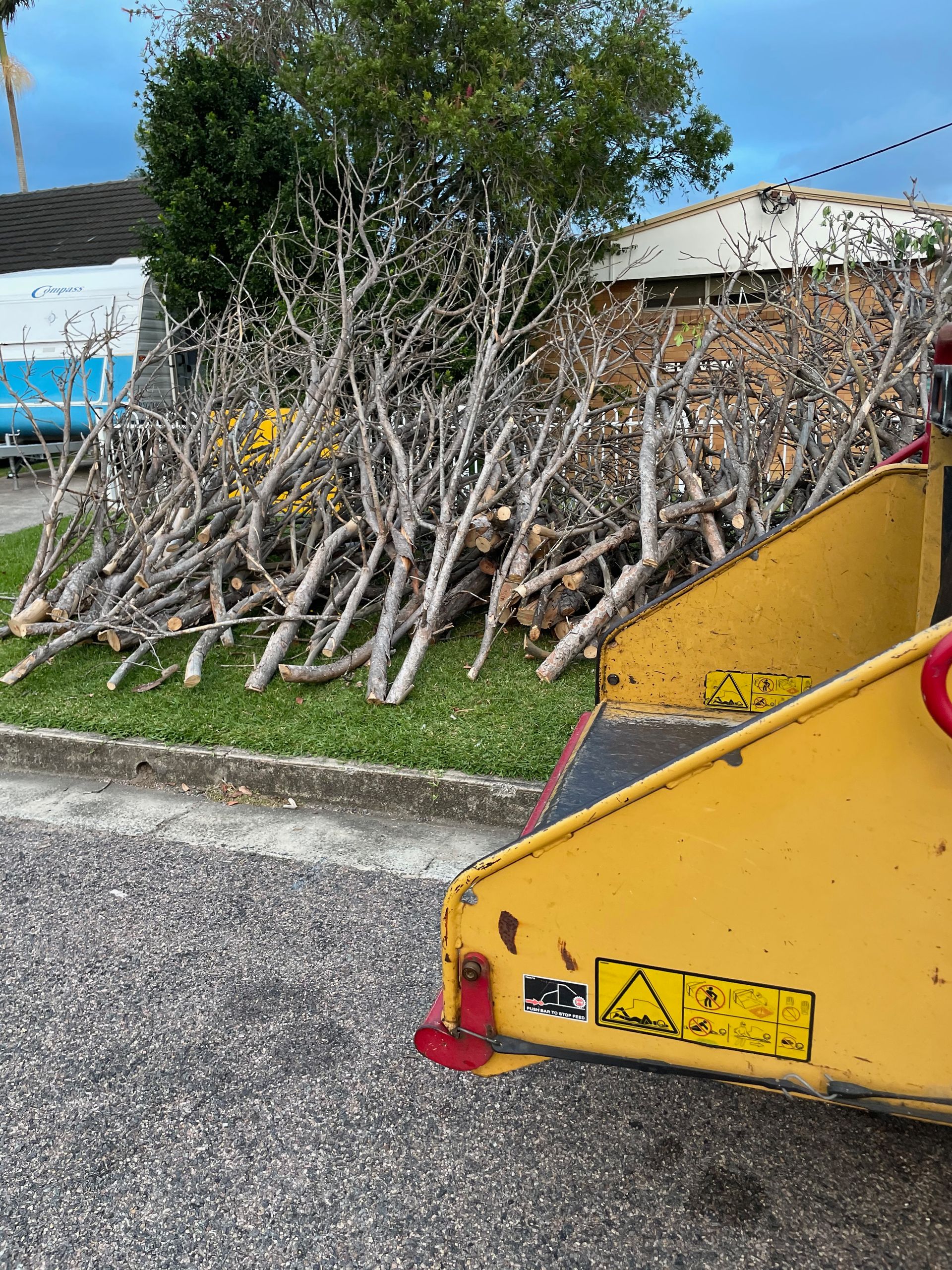 A pile of wood is sitting in front of a house with a yellow wood chipper machine next to it— Central Coast Tree Removal In Wyongah, NSW