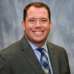A person wearing a gray suit, light blue checkered shirt, and a blue plaid tie, smiling against a mottled blue background.