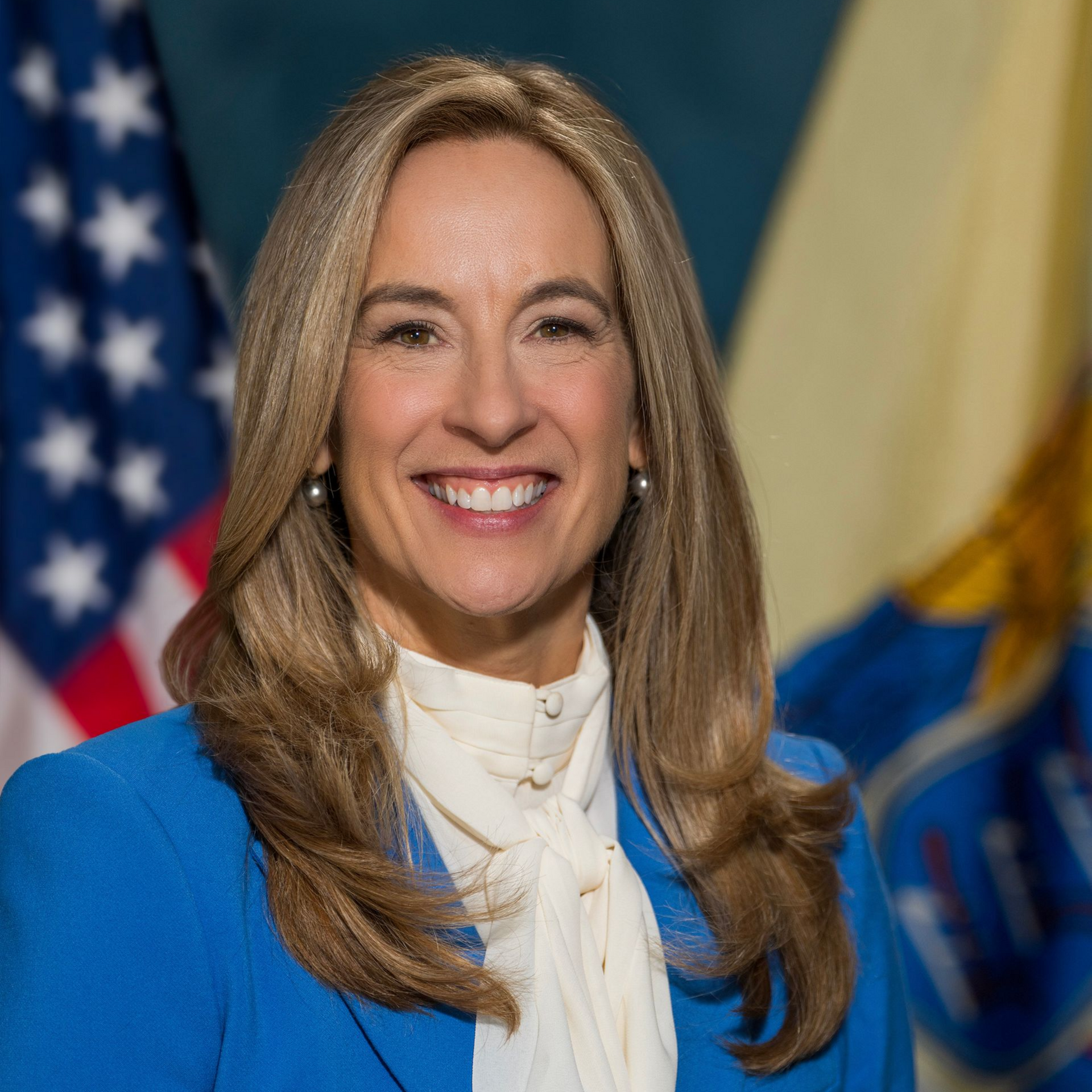 A woman smiling in a bright blue blazer and white blouse, with U.S. and state flags in the background.