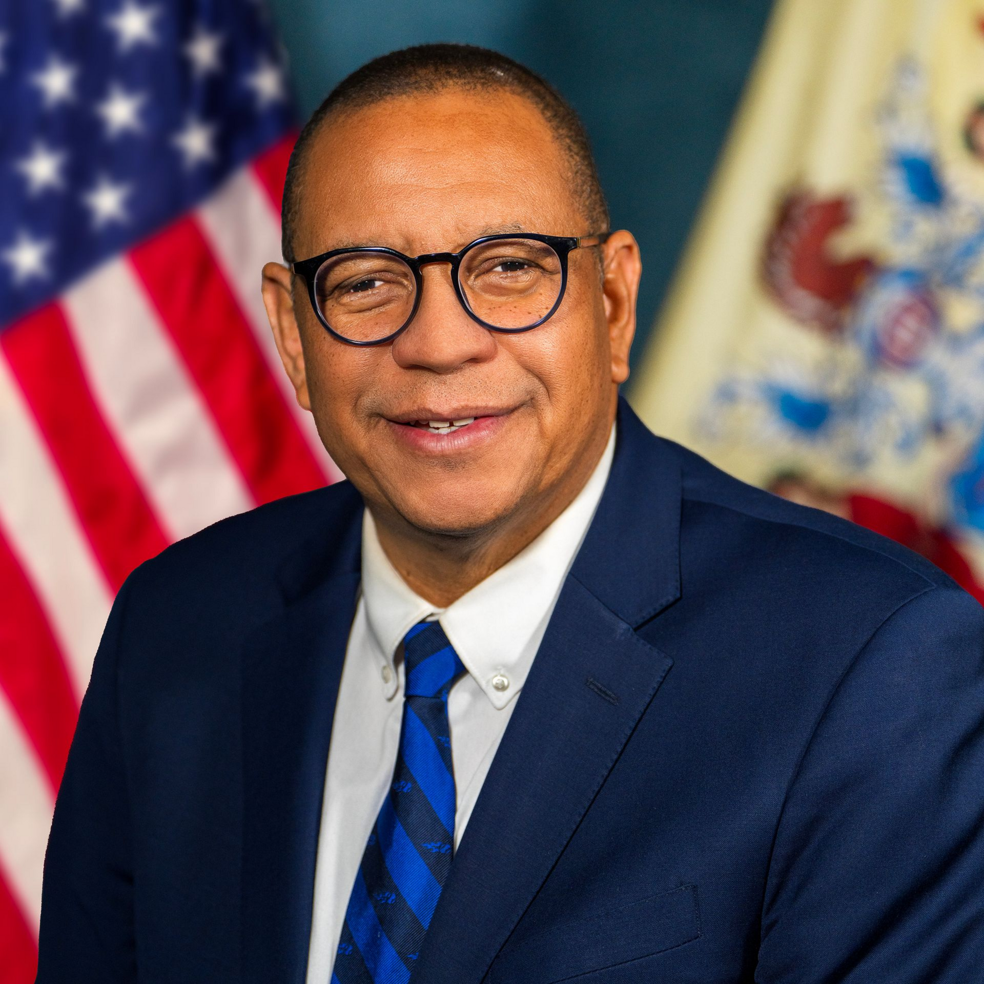 A man in a suit and blue tie wearing glasses smiles in front of U.S. and New Jersey state flags.
