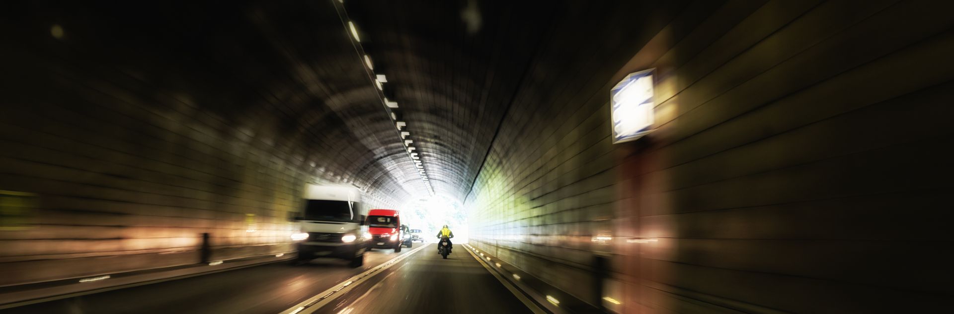 A motorcycle rides inside a dimly lit tunnel toward a bright exit, with vehicles ahead on the road.