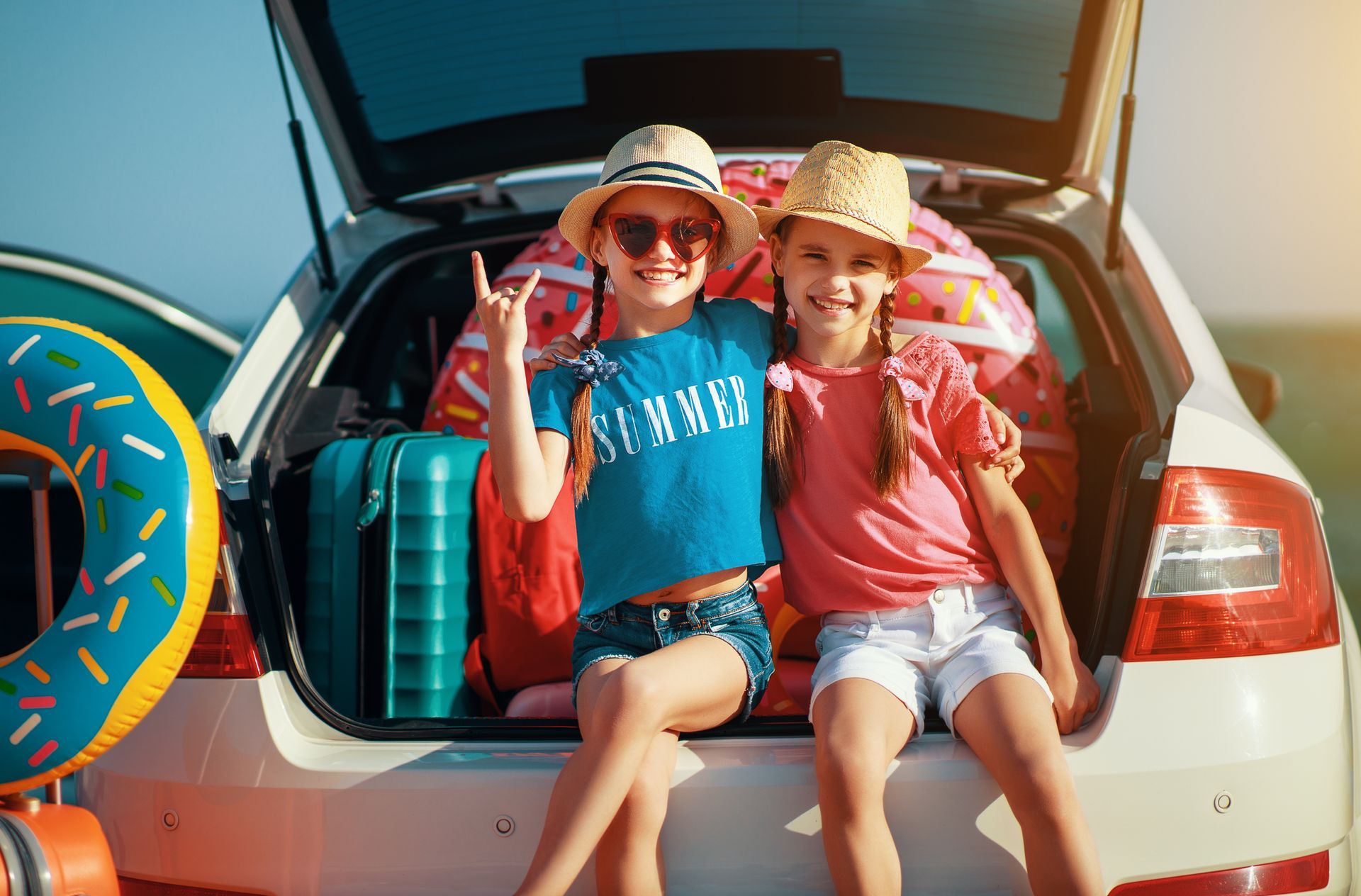 Two children in sun hats sit in the trunk of a car packed with luggage and pool floats, smiling at the camera.