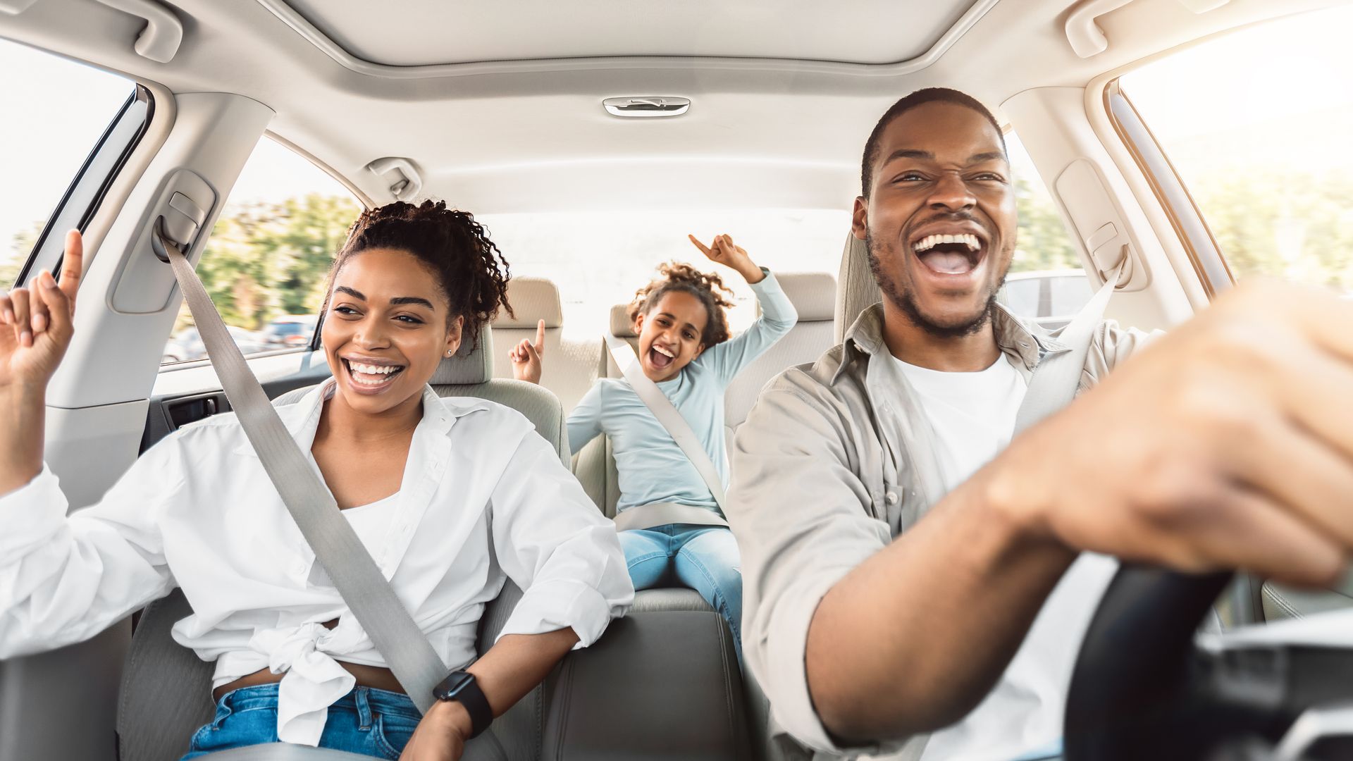 A family of three smiling and gesturing excitedly while riding in a car, with an adult driving.