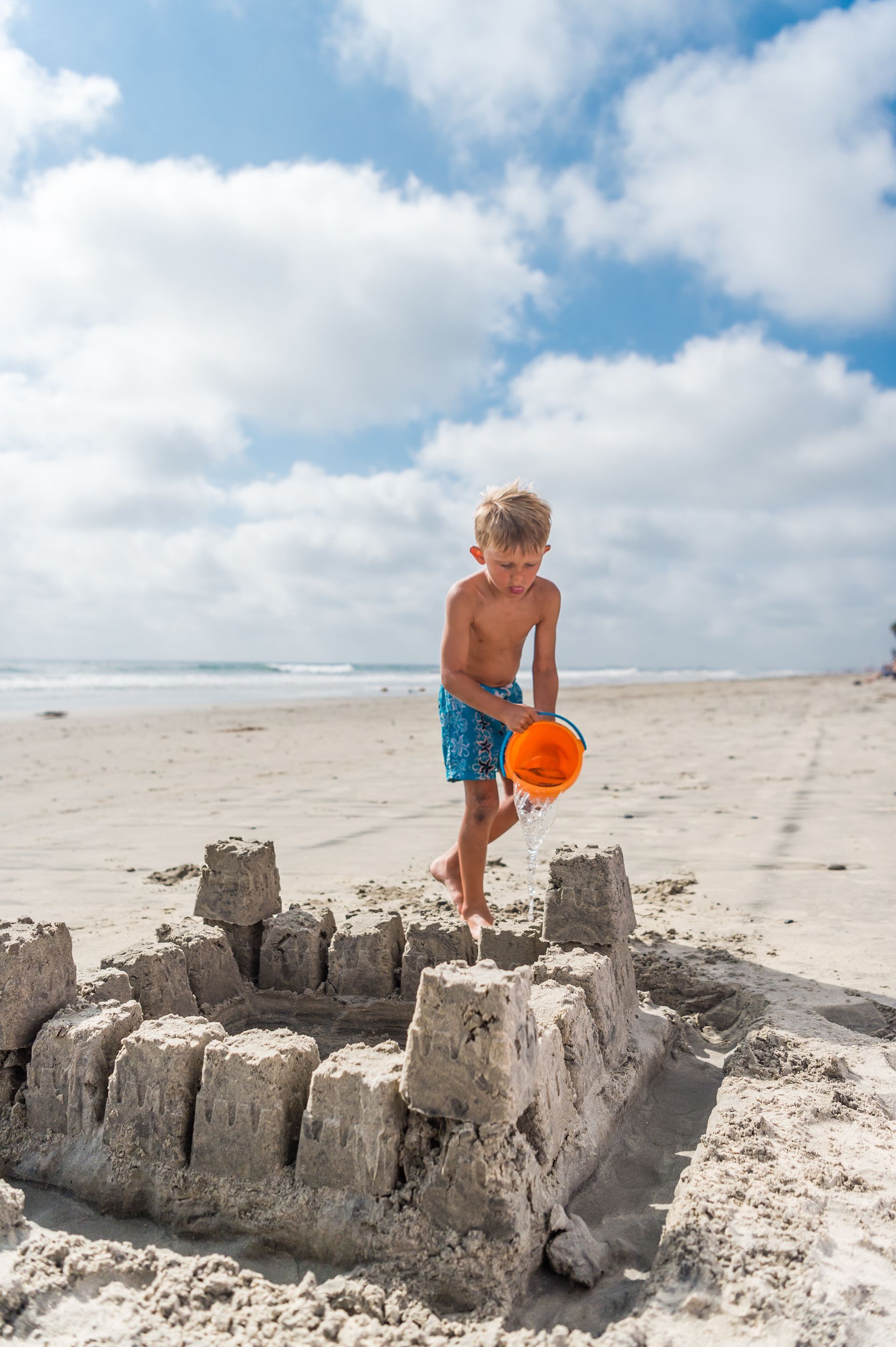 A child in blue swim trunks pours water from an orange bucket onto a sandcastle on a sunny beach.