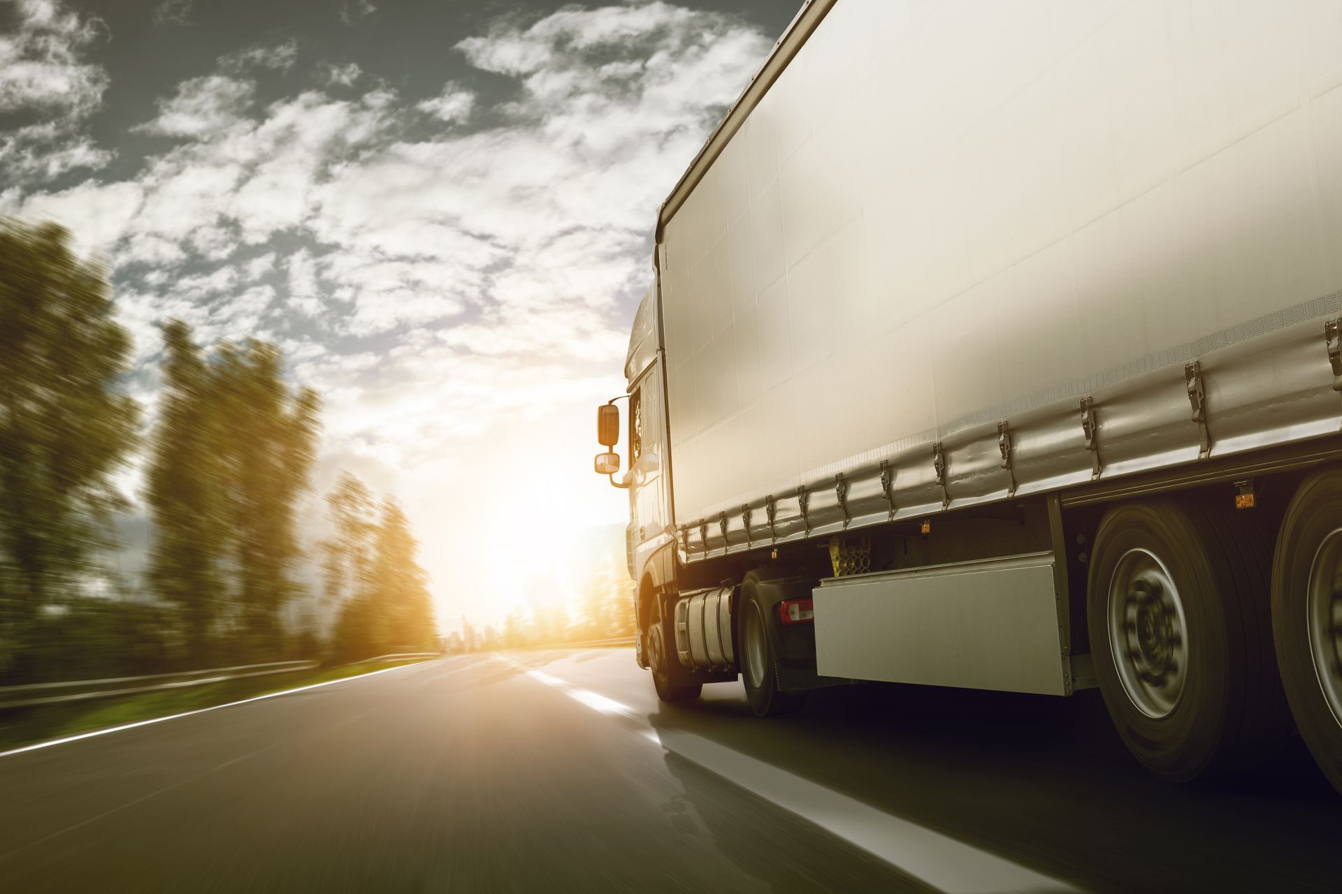 A white semi-truck drives on a road during a sunny day with trees in the background.