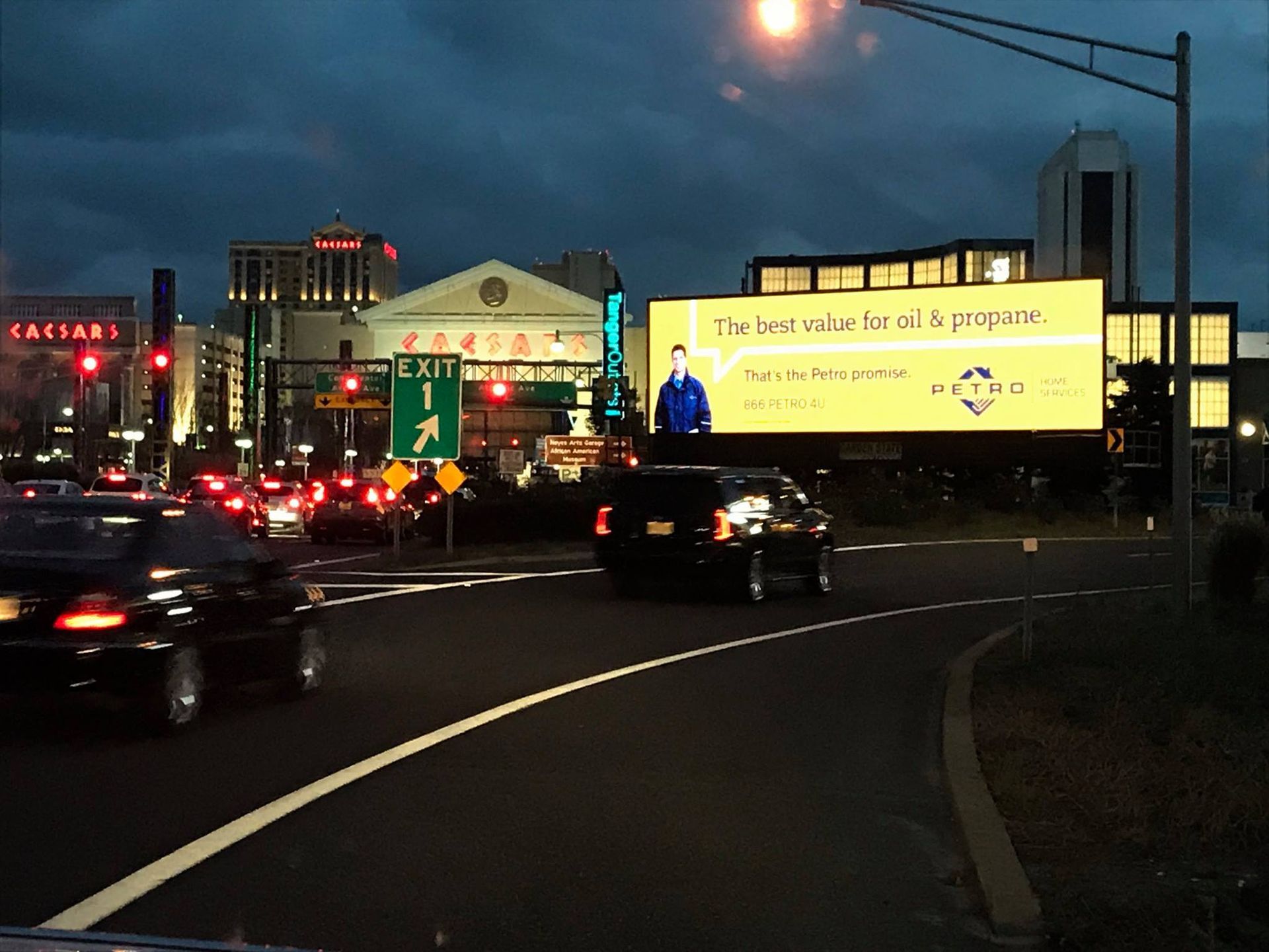 Cars driving on a road at dusk towards an exit sign and a large, bright digital billboard advertising energy services.