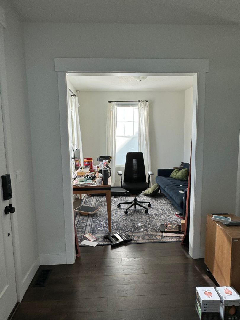 Doorway view into a cluttered living room with desk, black chair, patterned rug, and couch by a bright window