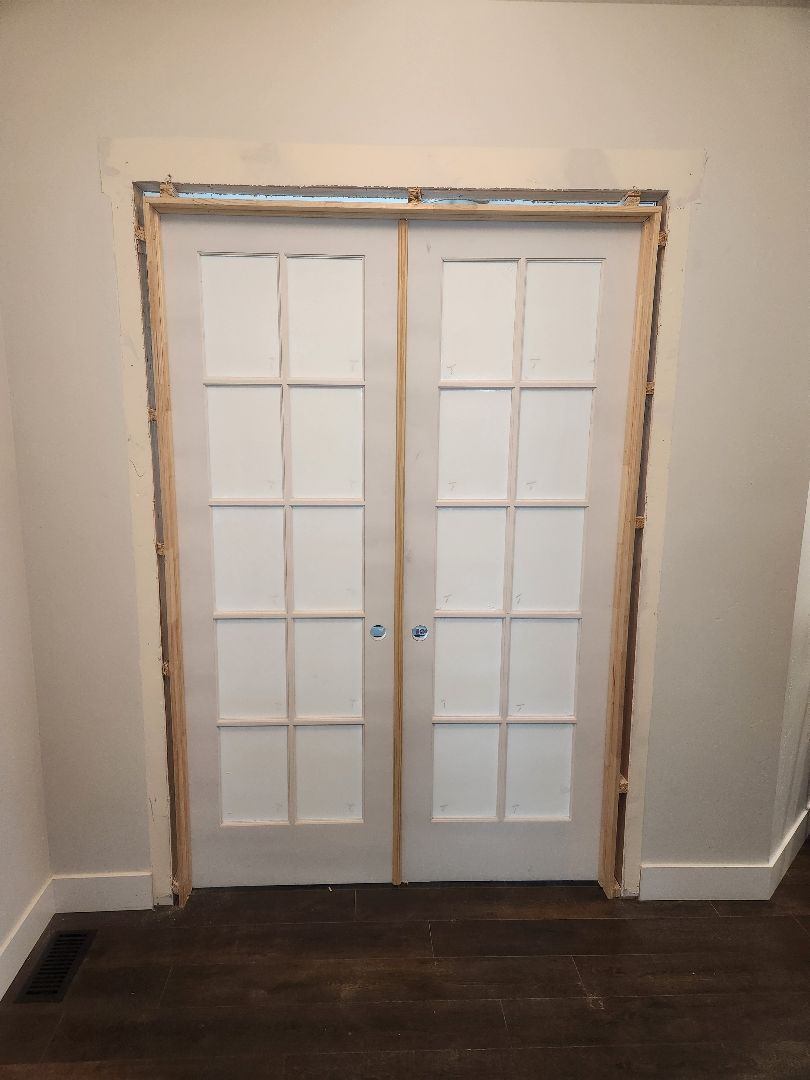 White French doors with glass panes in a plain room, framed by an unfinished doorway and dark wood floor