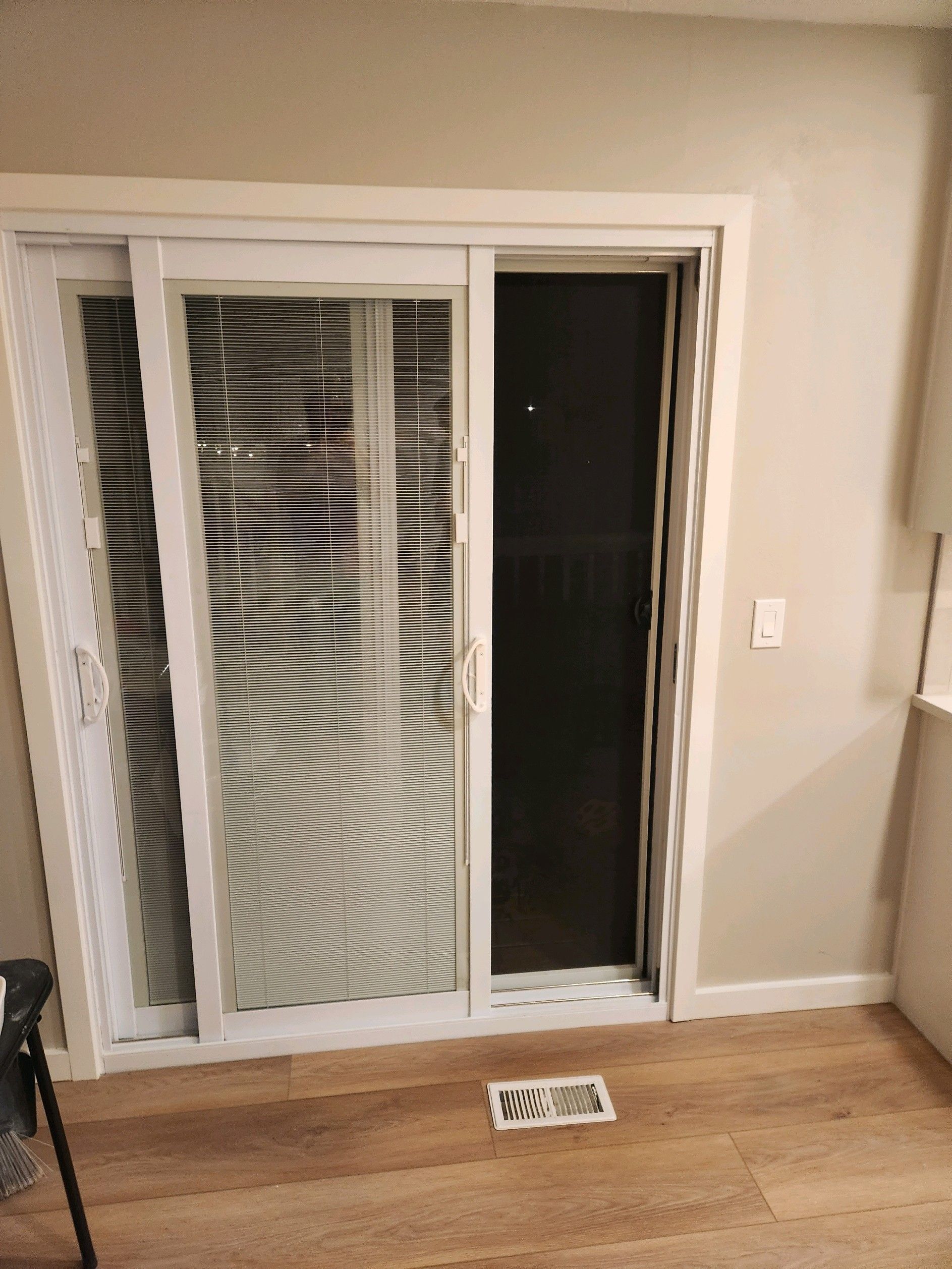 Sliding glass door with blinds, partly open to a dark patio, in a beige room with wood floor and small rug