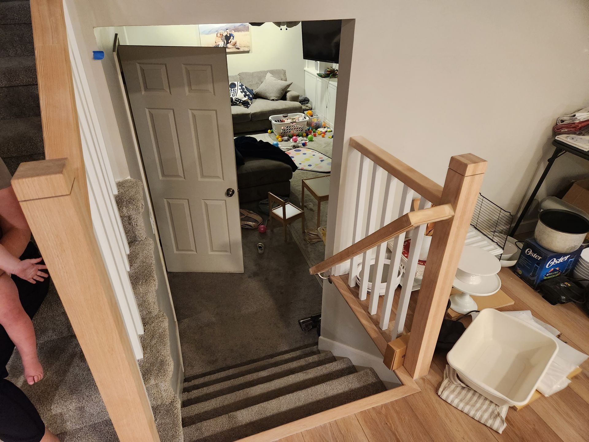 Stairwell opening to a cluttered room with a white door, wooden railing, and trash bin nearby