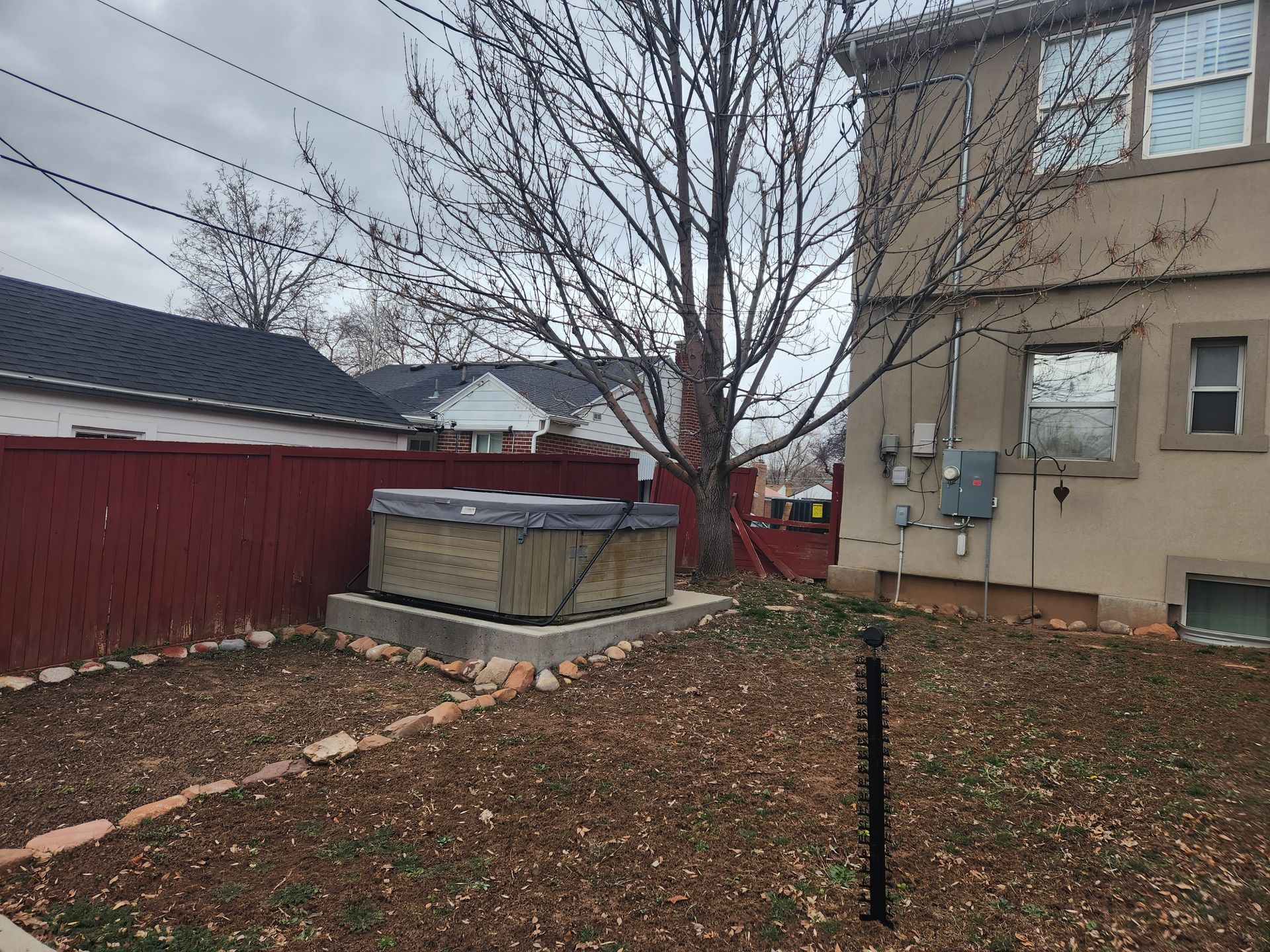 Backyard with leafless tree, red fence, hot tub, and beige house on an overcast day