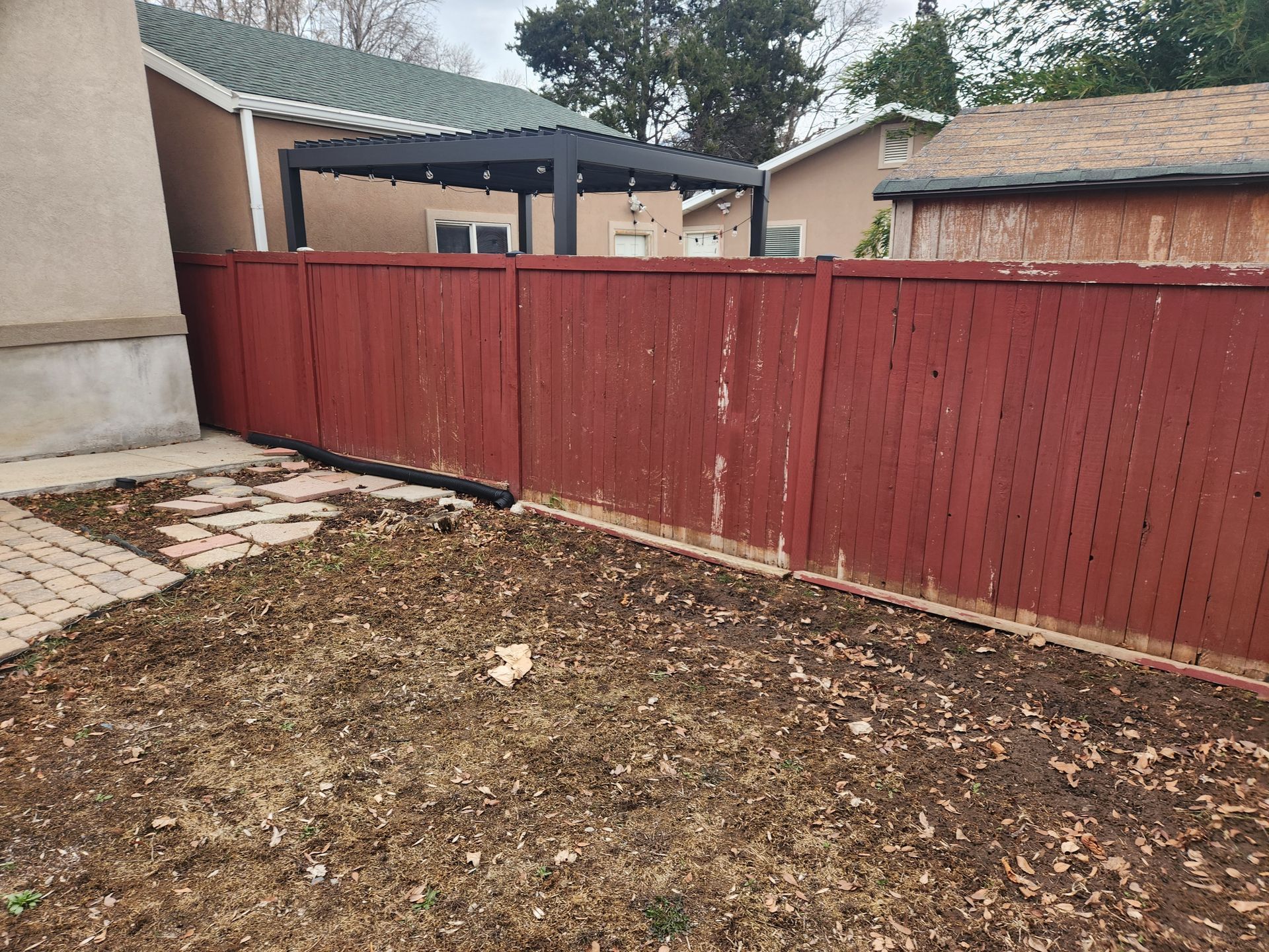 Backyard with red wooden fence, leaf-covered ground, and a covered patio beside a house