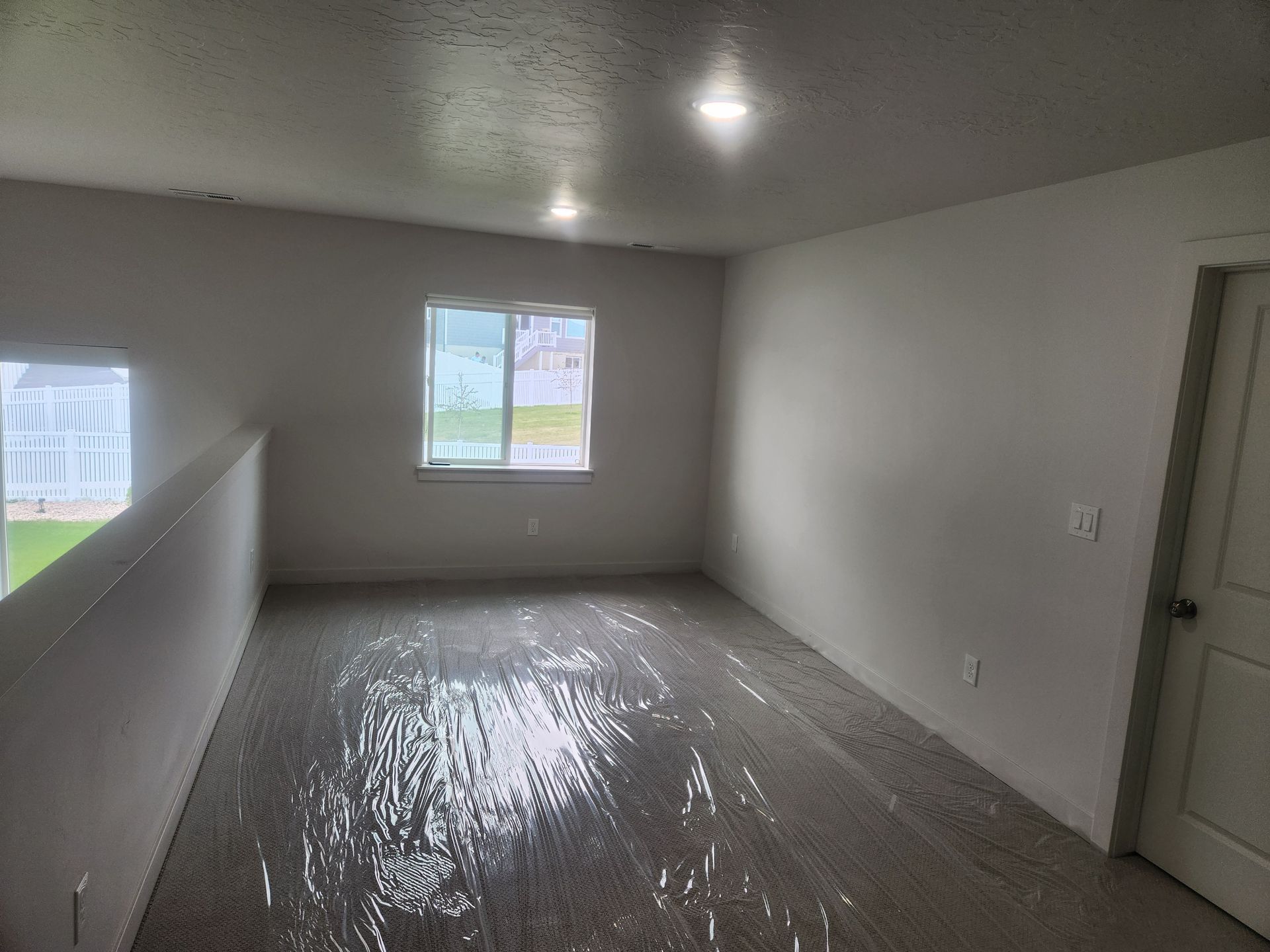 Empty unfurnished room with white walls, small windows, and a closed door, floor covered in plastic sheeting