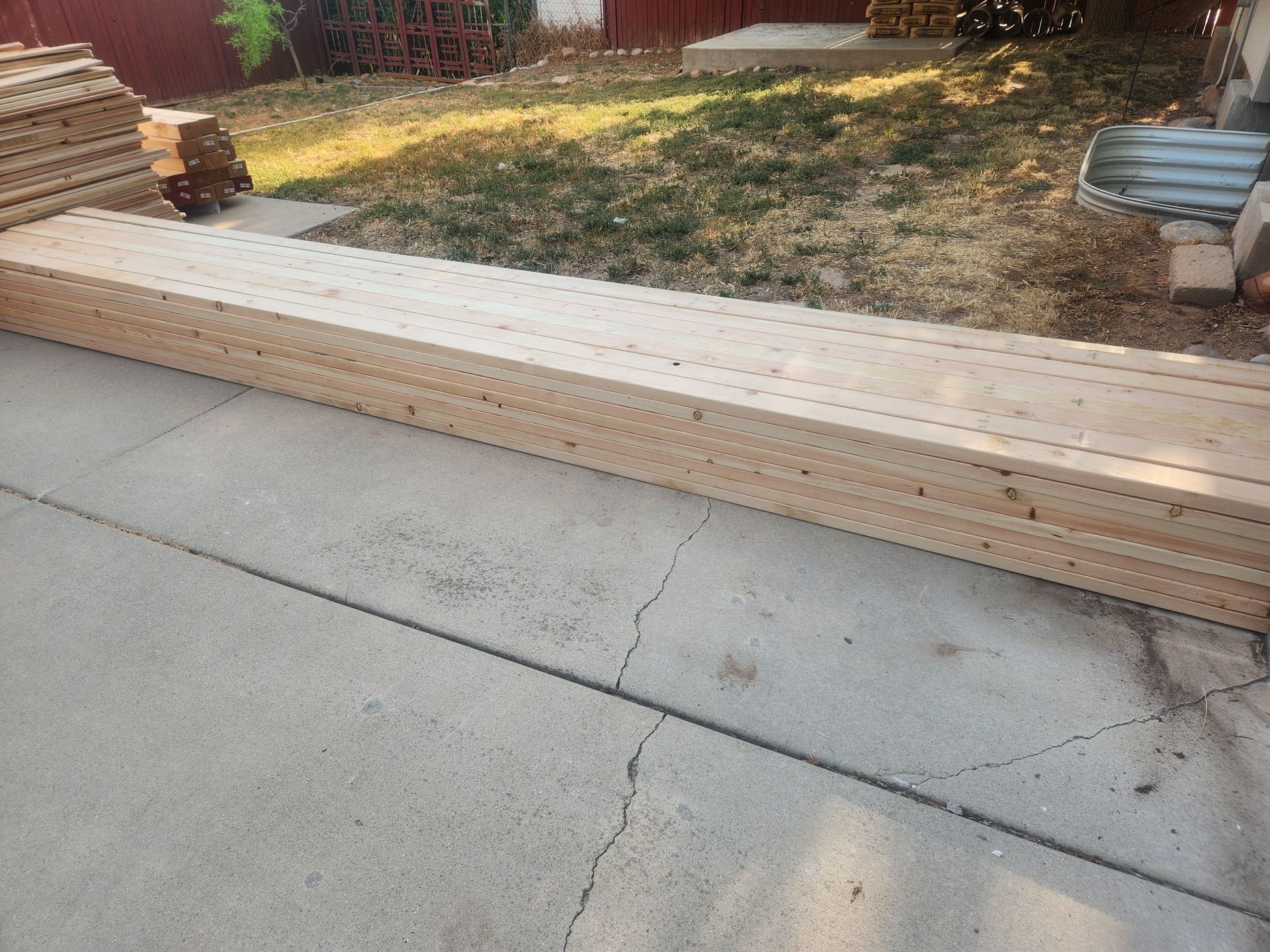 Long stack of lumber beside a driveway near a yard and house