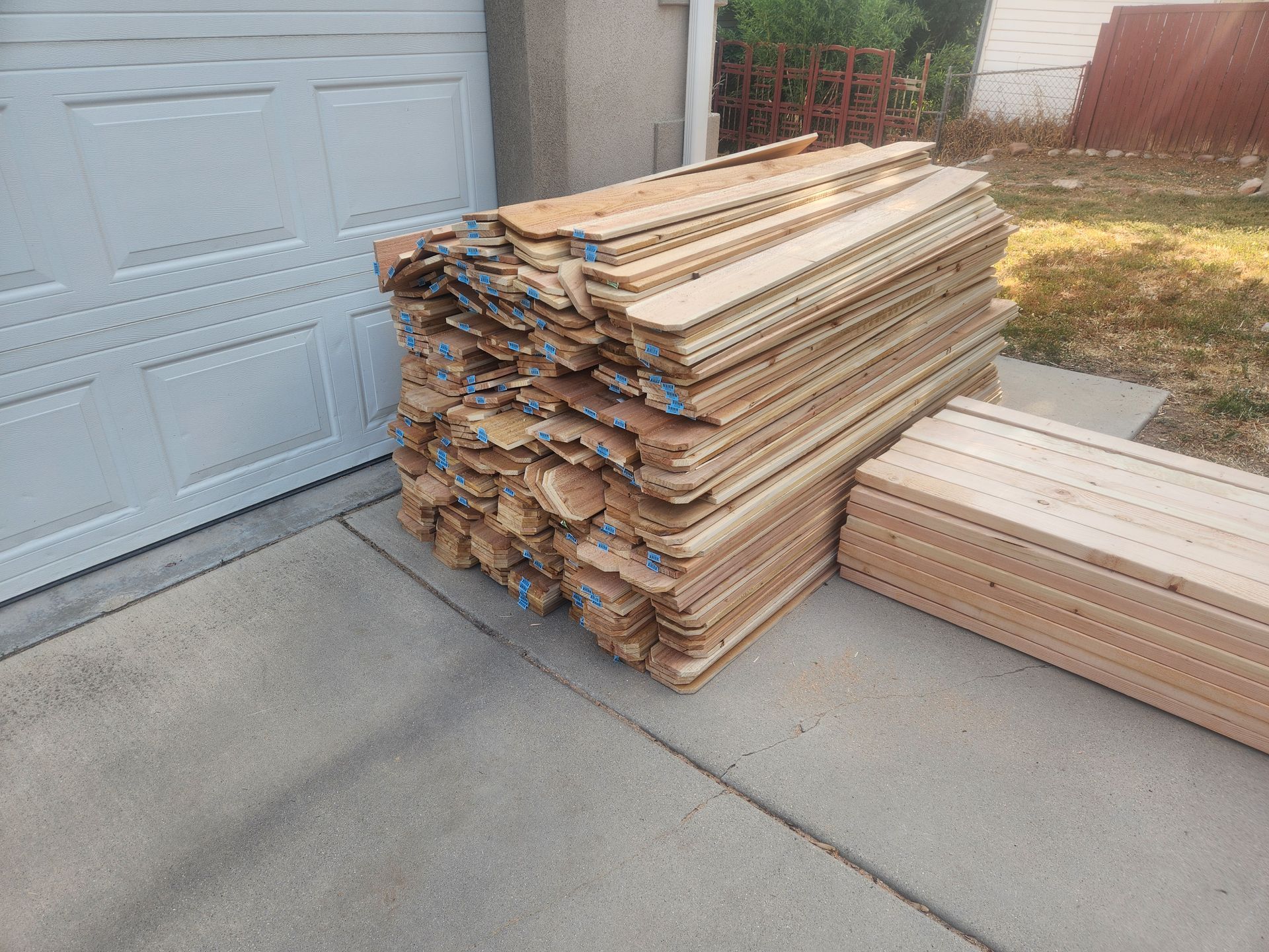 Stacks of lumber beside a garage on a concrete driveway, with some boards piled on the lawn.