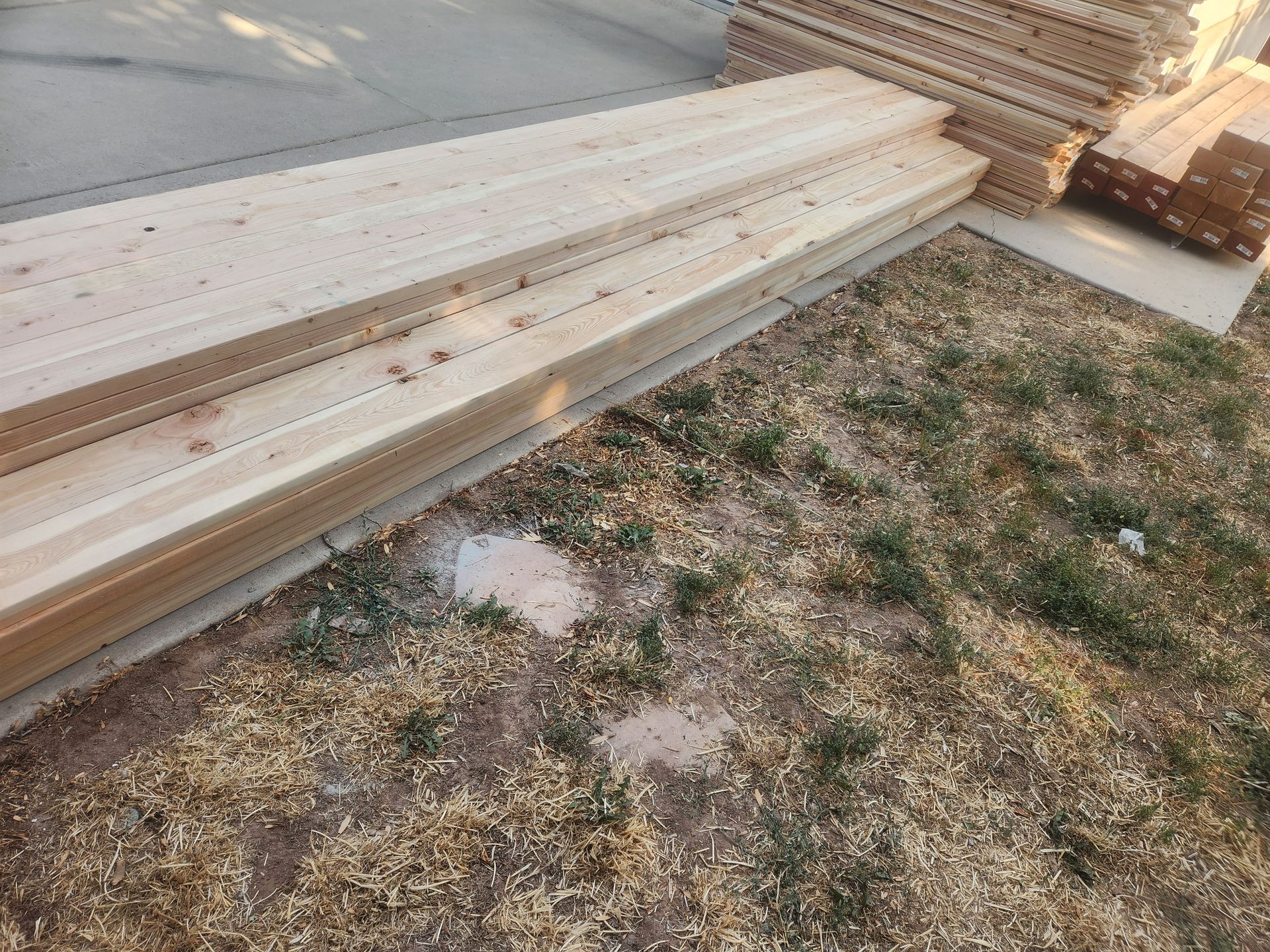 Stacks of light-colored lumber on grass beside a paved area, with more wood piled in the background.