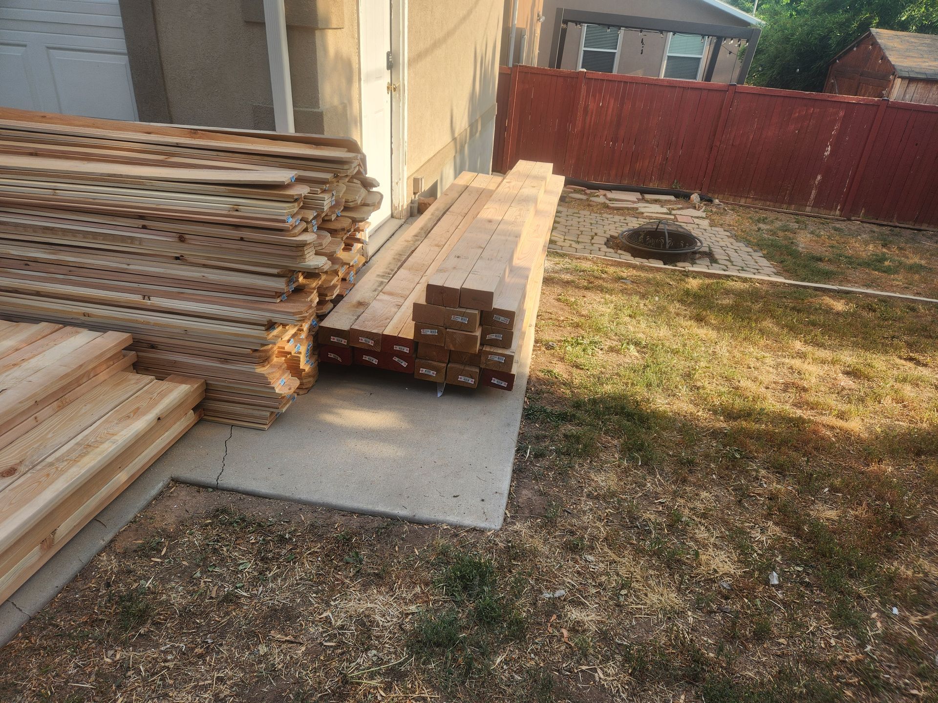 Stacks of lumber on concrete beside a red fence in a backyard.