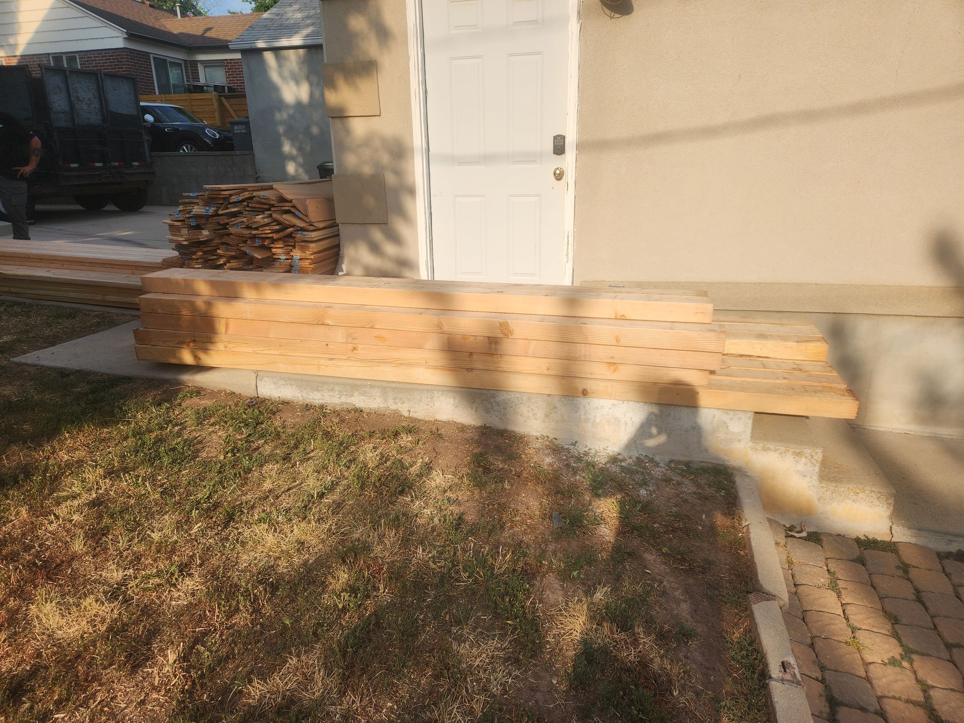 Stacked lumber and long wooden beams against a beige house in a sunny yard