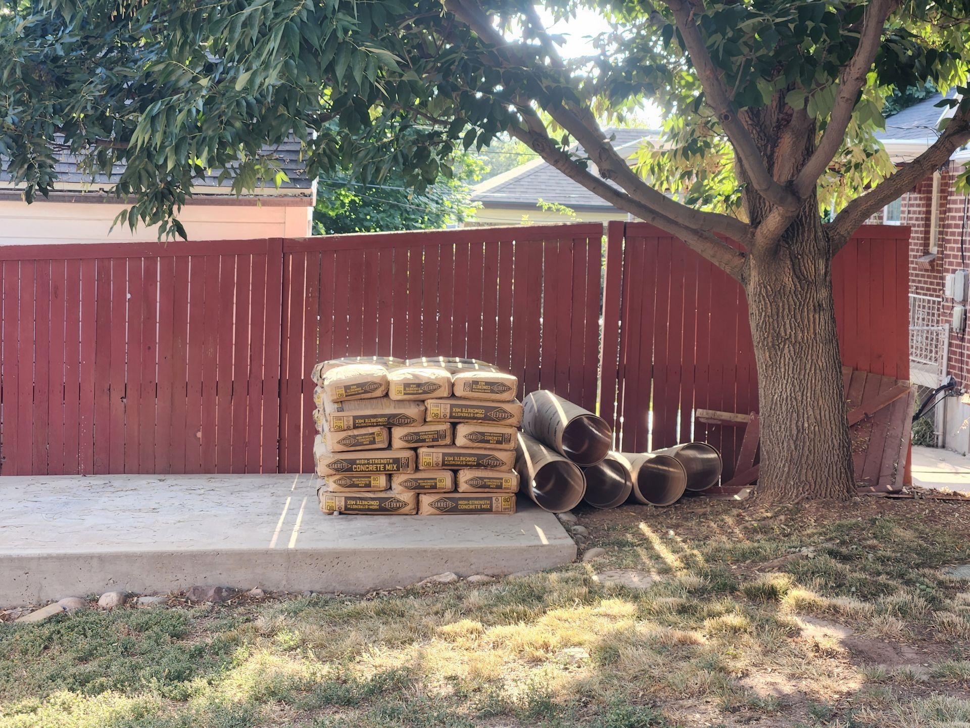 Stacks of cement bags beside a red fence under a tree in a yard, with logs nearby.