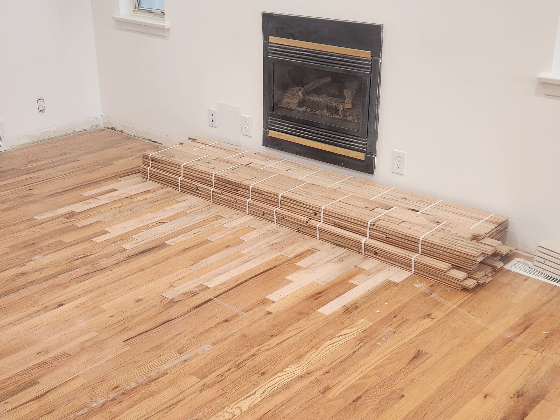 Living room with hardwood floor and a fireplace, with a long strip of flooring materials laid out for installation