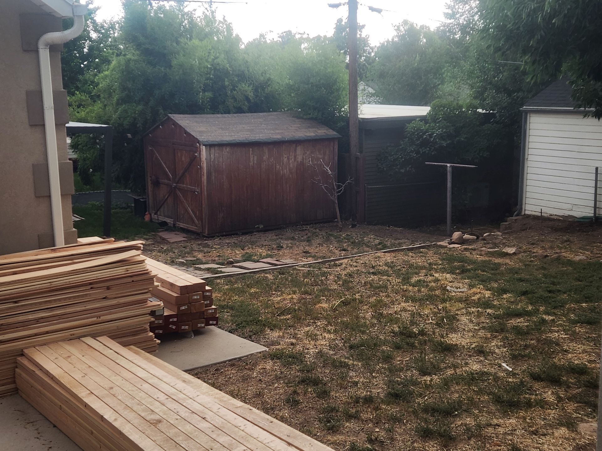 Backyard with stacked lumber, a wooden shed, and a patchy grass yard near a fence