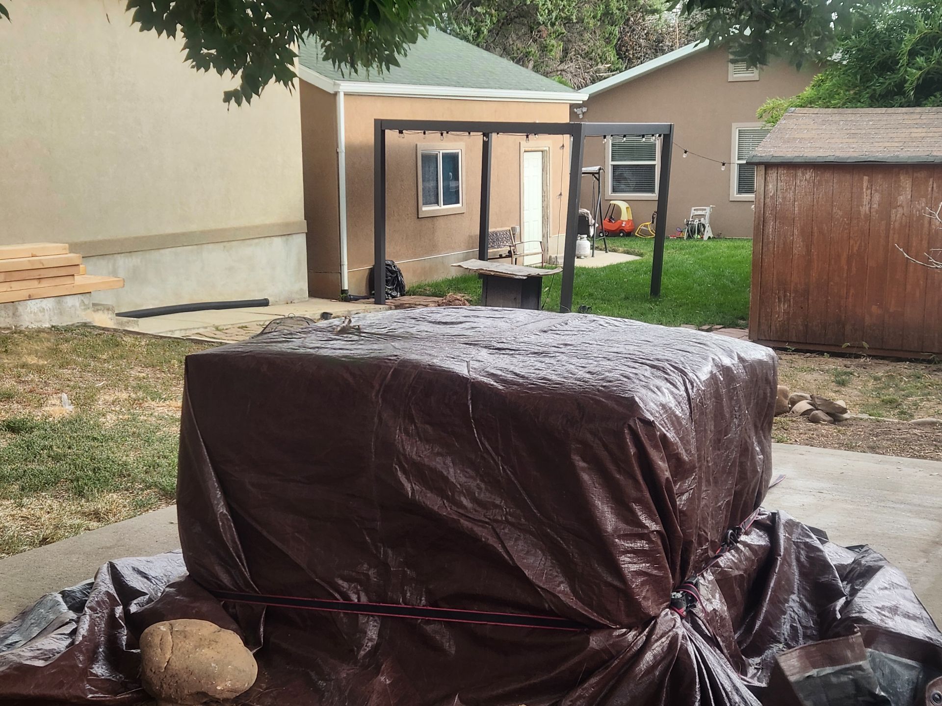 Covered backyard object wrapped in brown tarp beside a shed and patio.