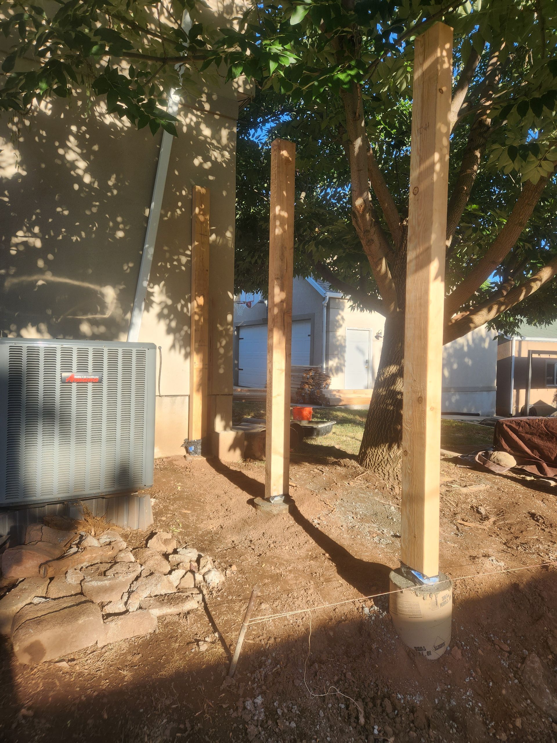 Backyard with wooden posts, an outdoor AC unit, and trees casting shade over dirt ground.