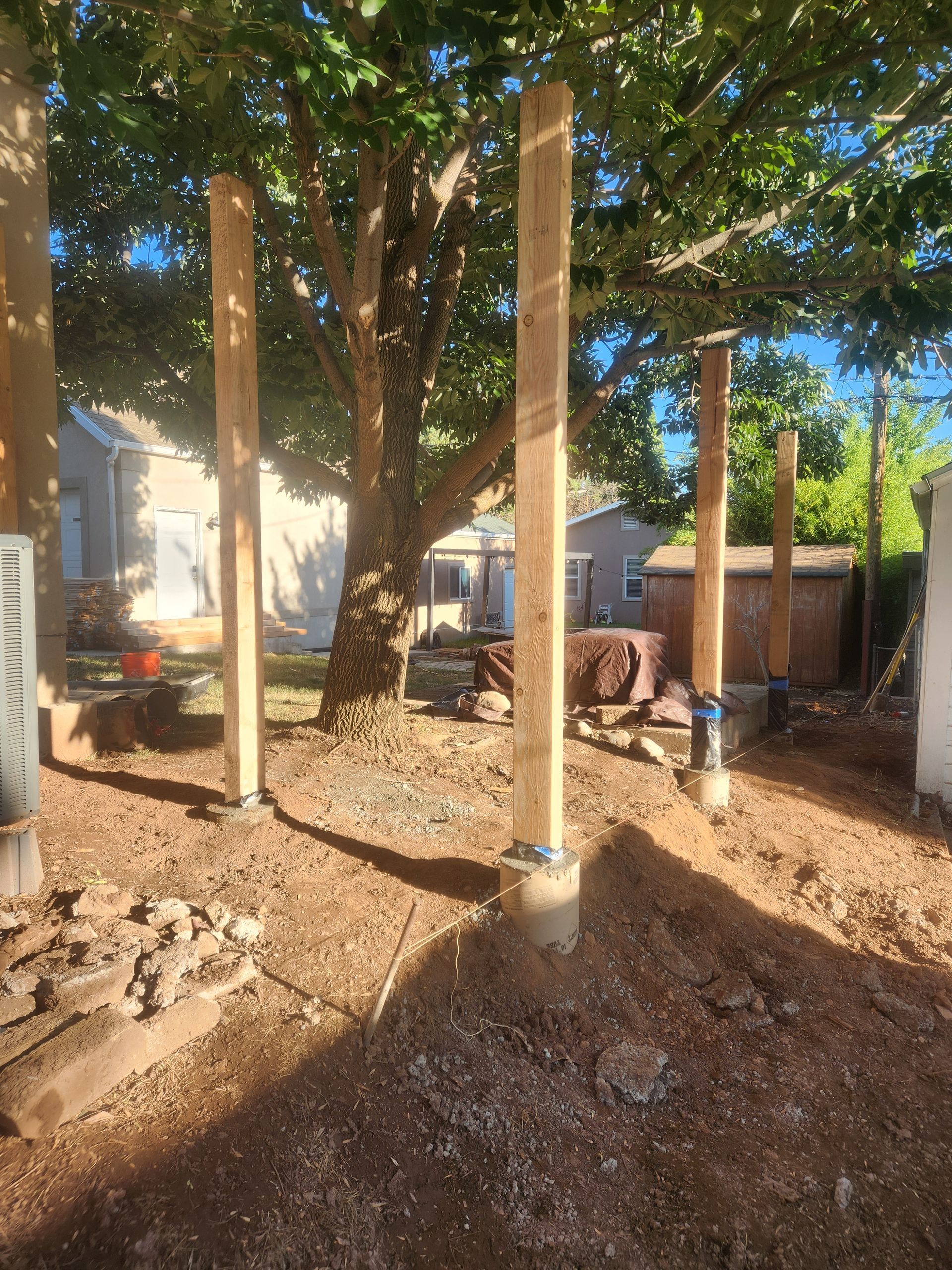 Backyard construction site with several upright wooden posts set in dirt and gravel near trees and a fence